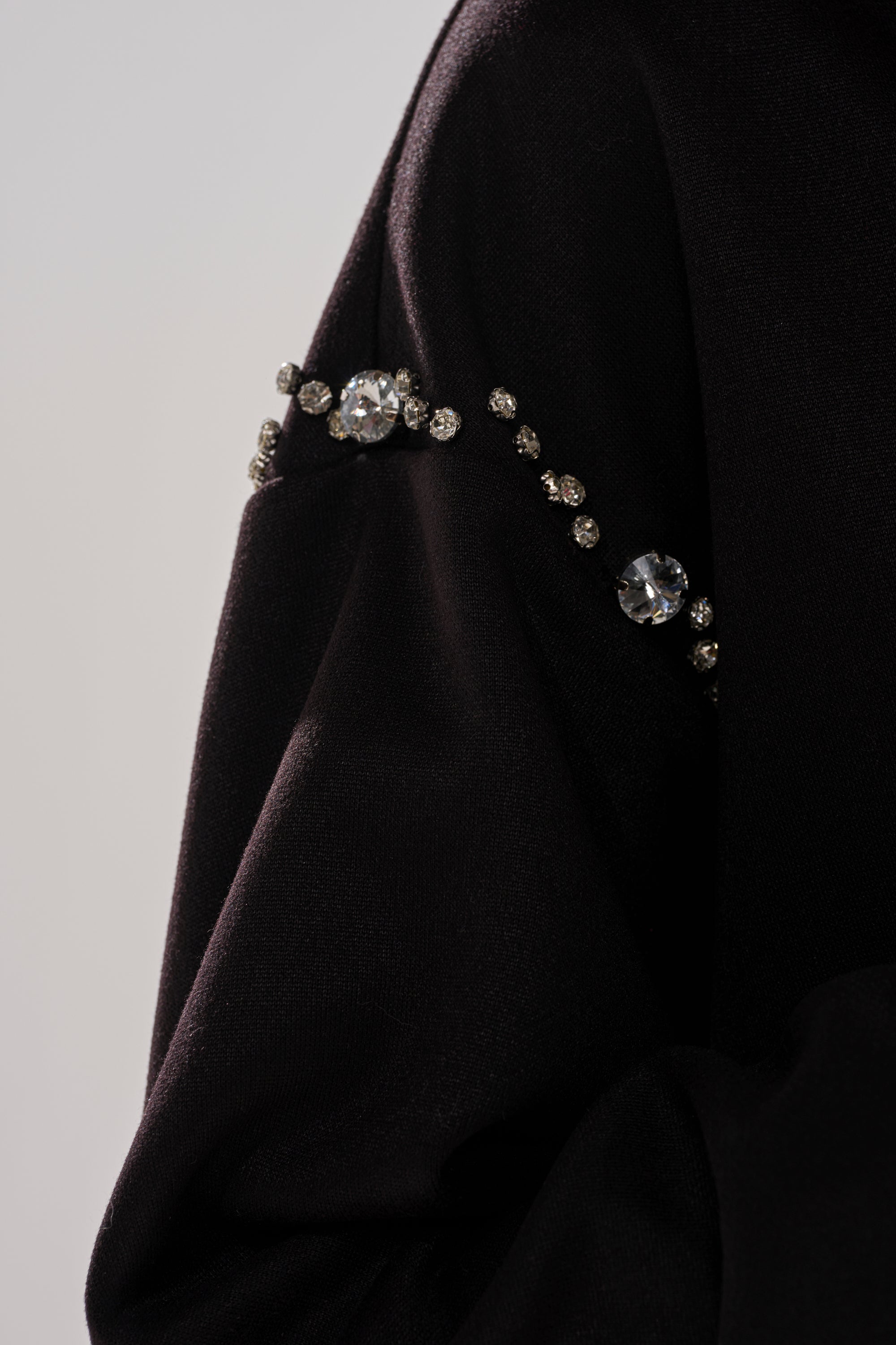 Close-up of the COZY HOLIDAY RHINESTONE ZIP UP SWEATSHIRT IN BLACK, featuring a black sleeve embellished with a curved line of sparkling rhinestones and a few larger gems, creating an elegant detail against a plain, light background.