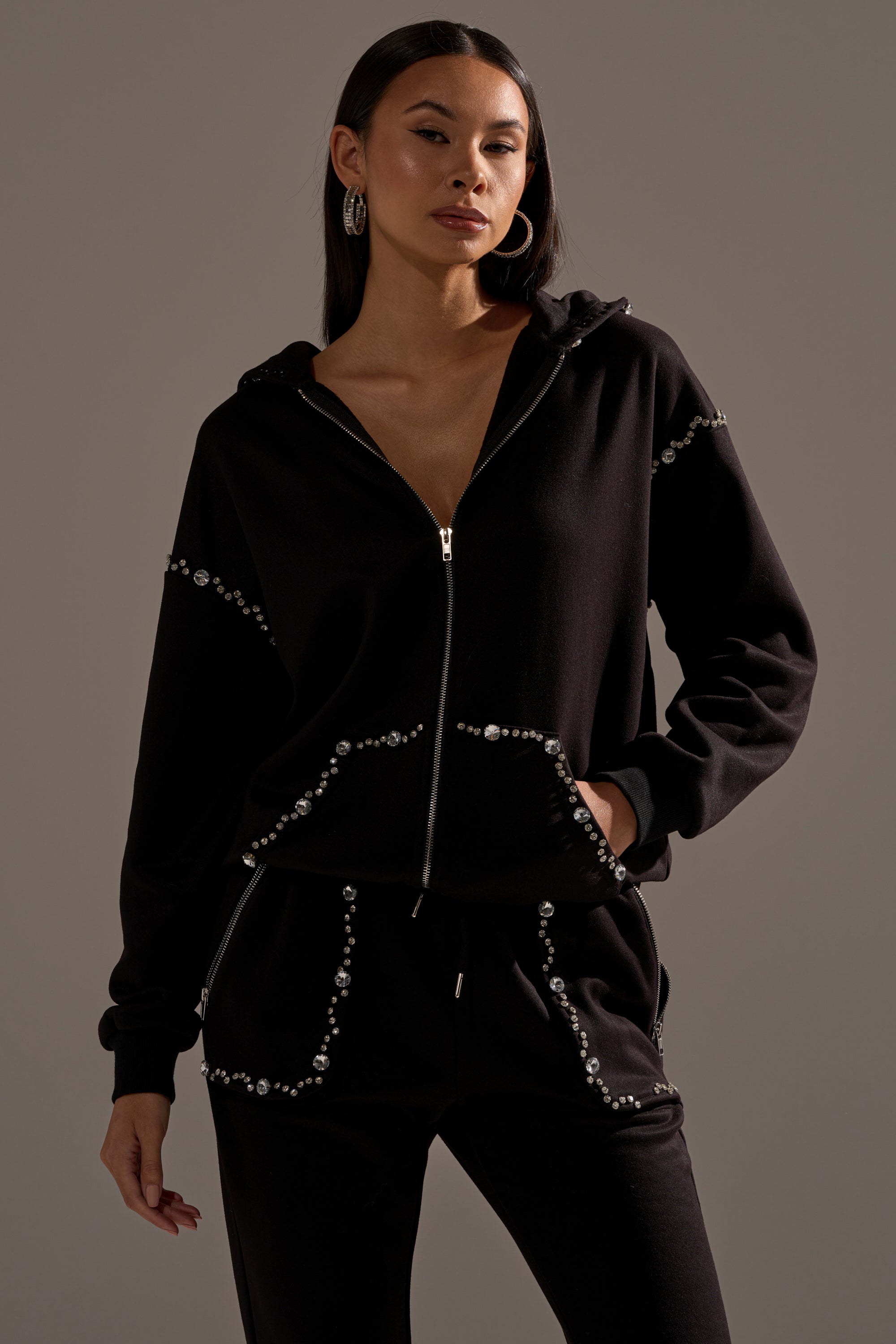A woman wearing the COZY HOLIDAY RHINESTONE ZIP UP SWEATSHIRT IN BLACK and matching pants stands with one hand in her pocket against a plain background.