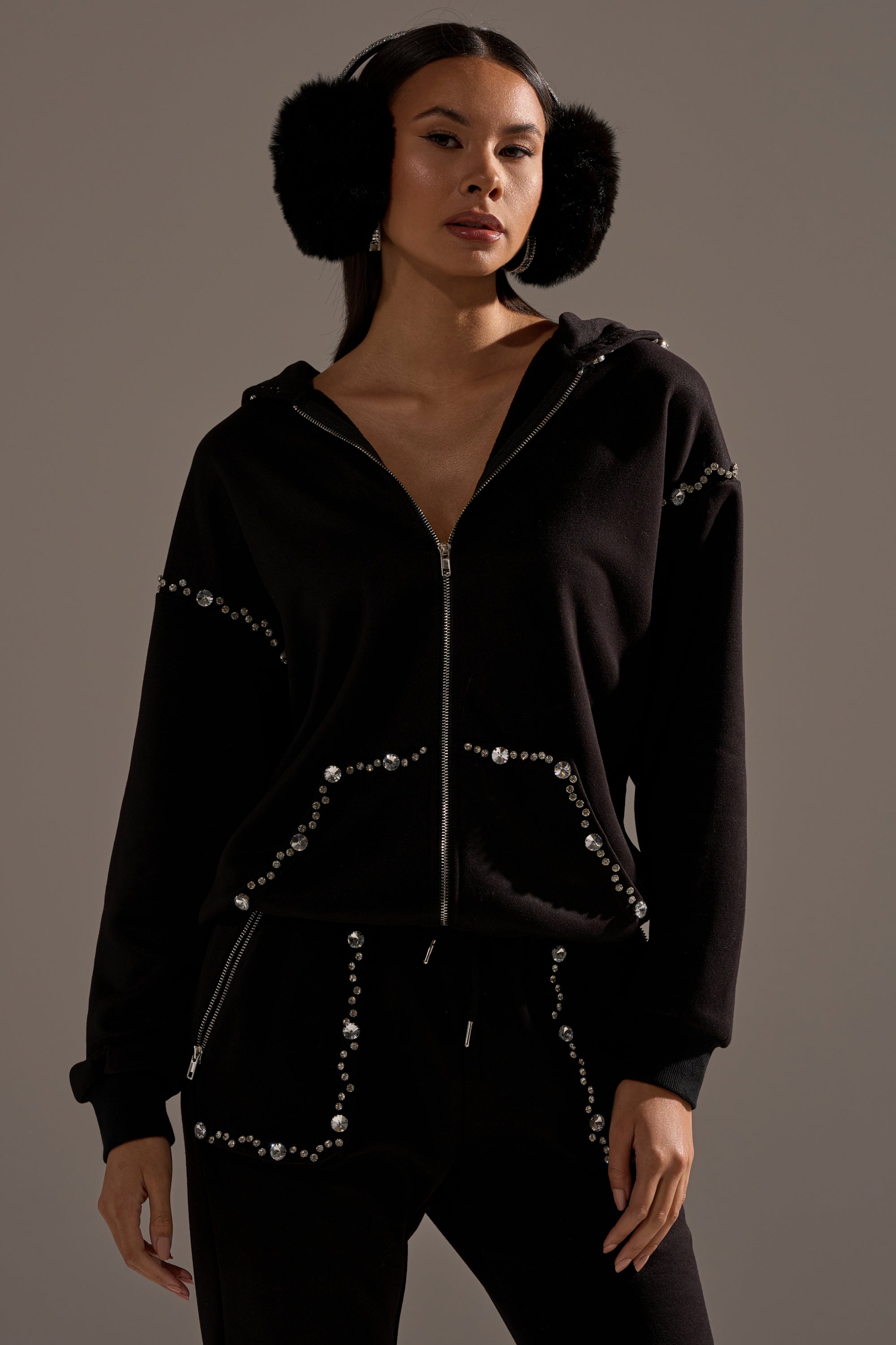 A woman with long dark hair and black earmuffs stands confidently against a neutral background, wearing the COZY HOLIDAY RHINESTONE ZIP UP SWEATSHIRT IN BLACK and looking slightly past the camera.