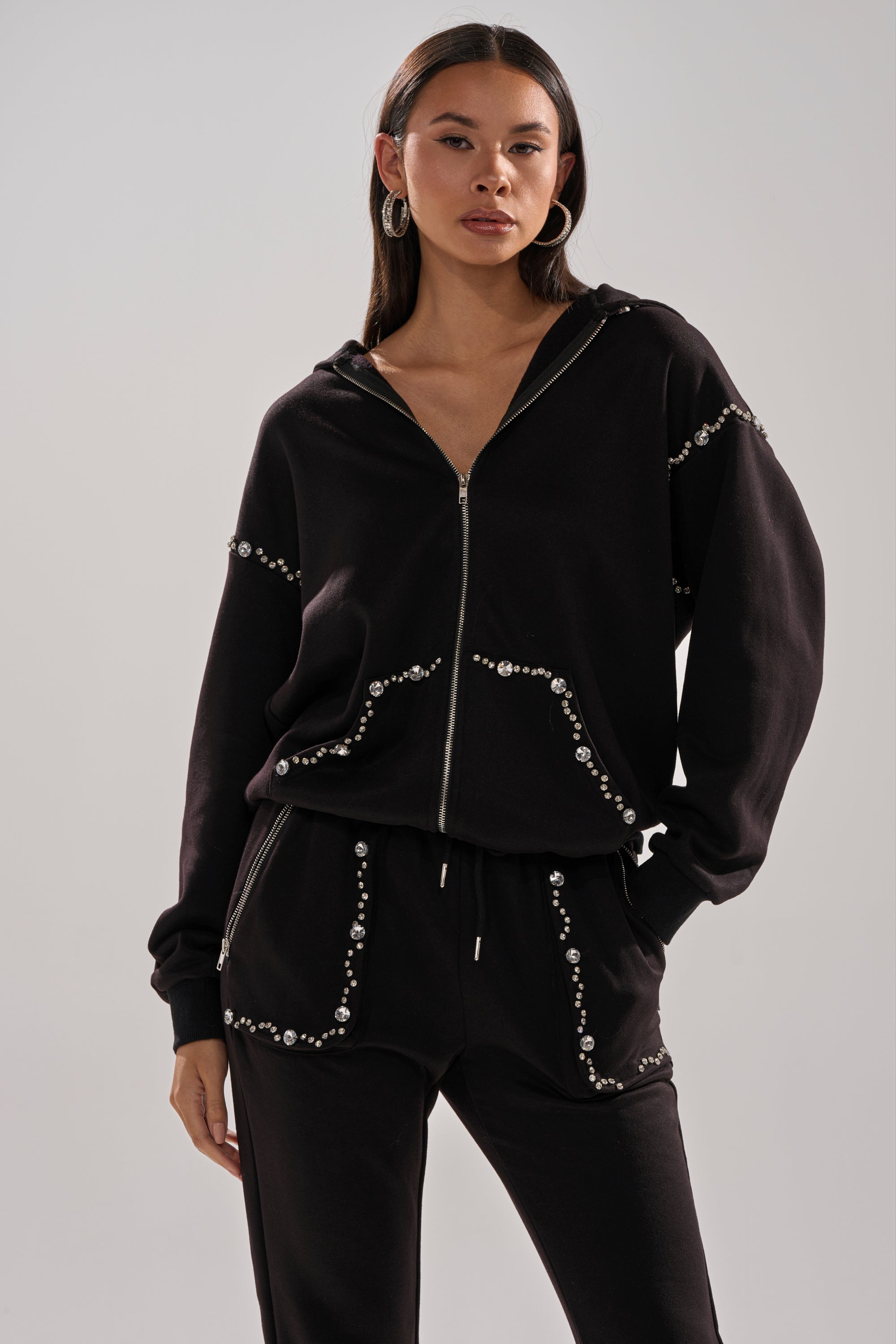 A woman confidently poses with one hand in her pocket, wearing the COZY HOLIDAY RHINESTONE ZIP UP SWEATSHIRT IN BLACK, adorned with silver studs and paired with matching pants.