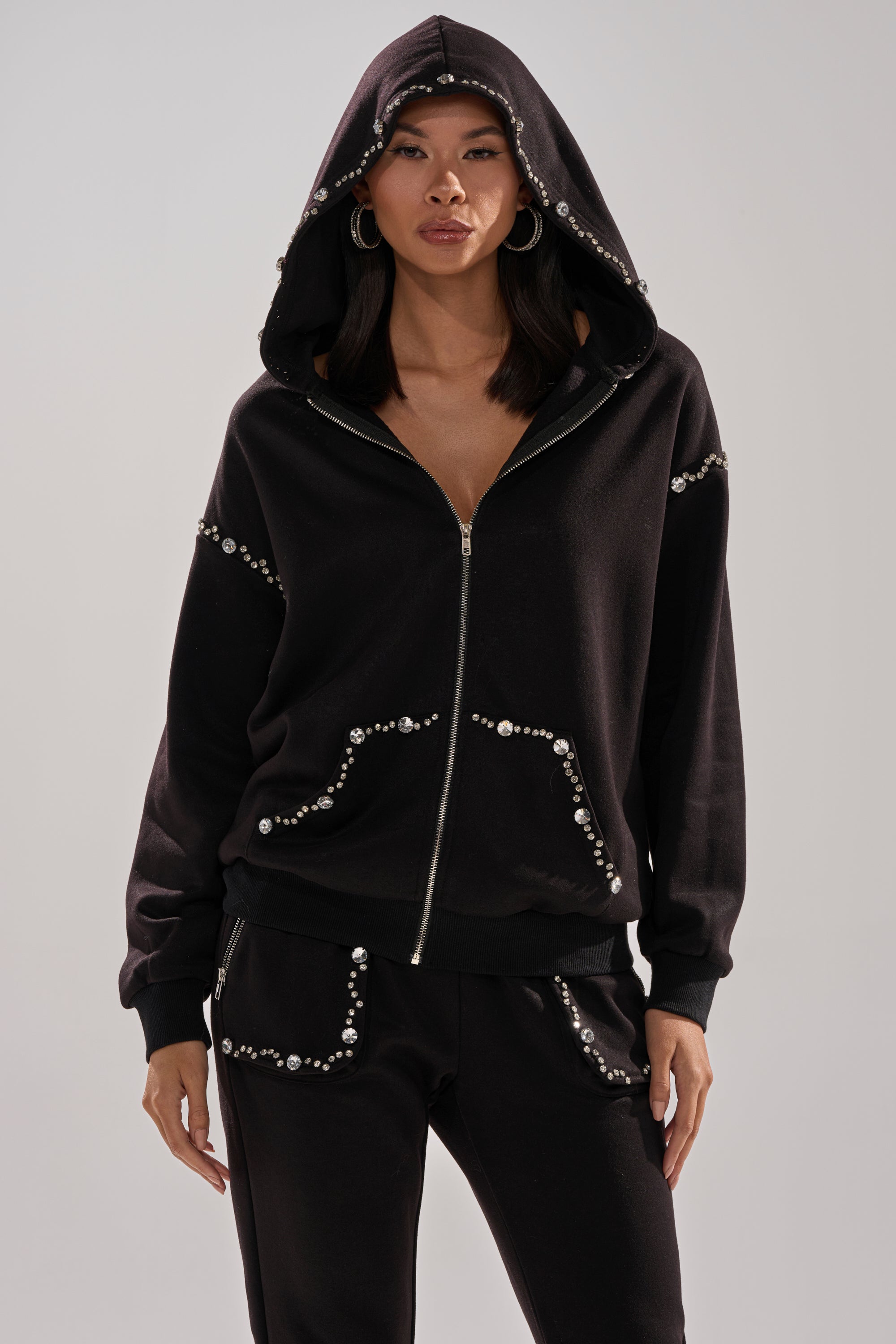 A woman models the COZY HOLIDAY RHINESTONE ZIP UP SWEATSHIRT IN BLACK with its hood up and silver rhinestone details, paired with matching black pants, against a light gray background.