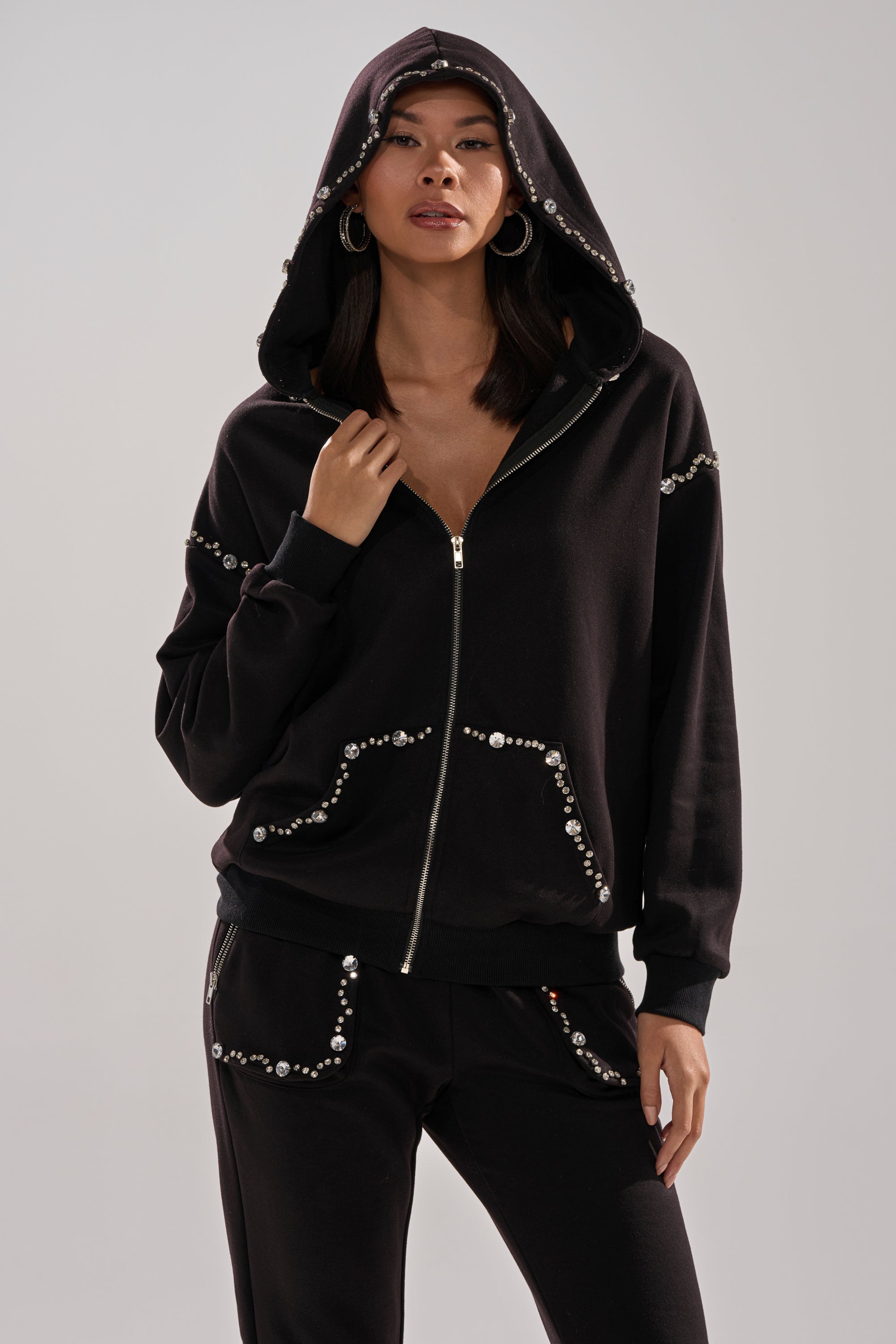 A woman wearing the AKIRA Label COZY HOLIDAY RHINESTONE ZIP UP SWEATSHIRT IN BLACK with matching pants stands confidently, one hand on her collar, against a plain background.