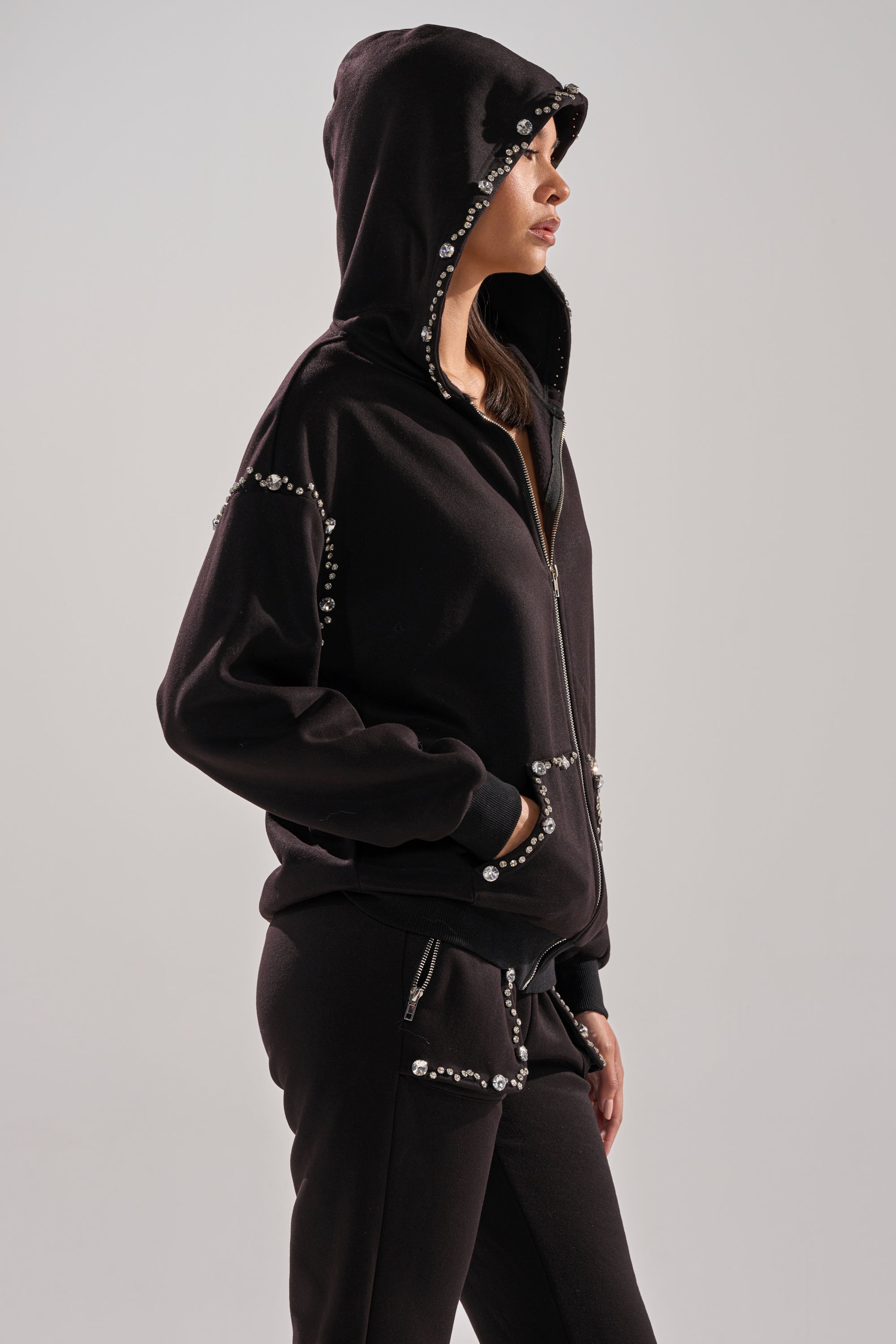 A person stands in profile with hands in pockets, wearing the COZY HOLIDAY RHINESTONE ZIP UP SWEATSHIRT IN BLACK from AKIRA Label, featuring silver studs. The hood is up and the background is plain and light-colored.