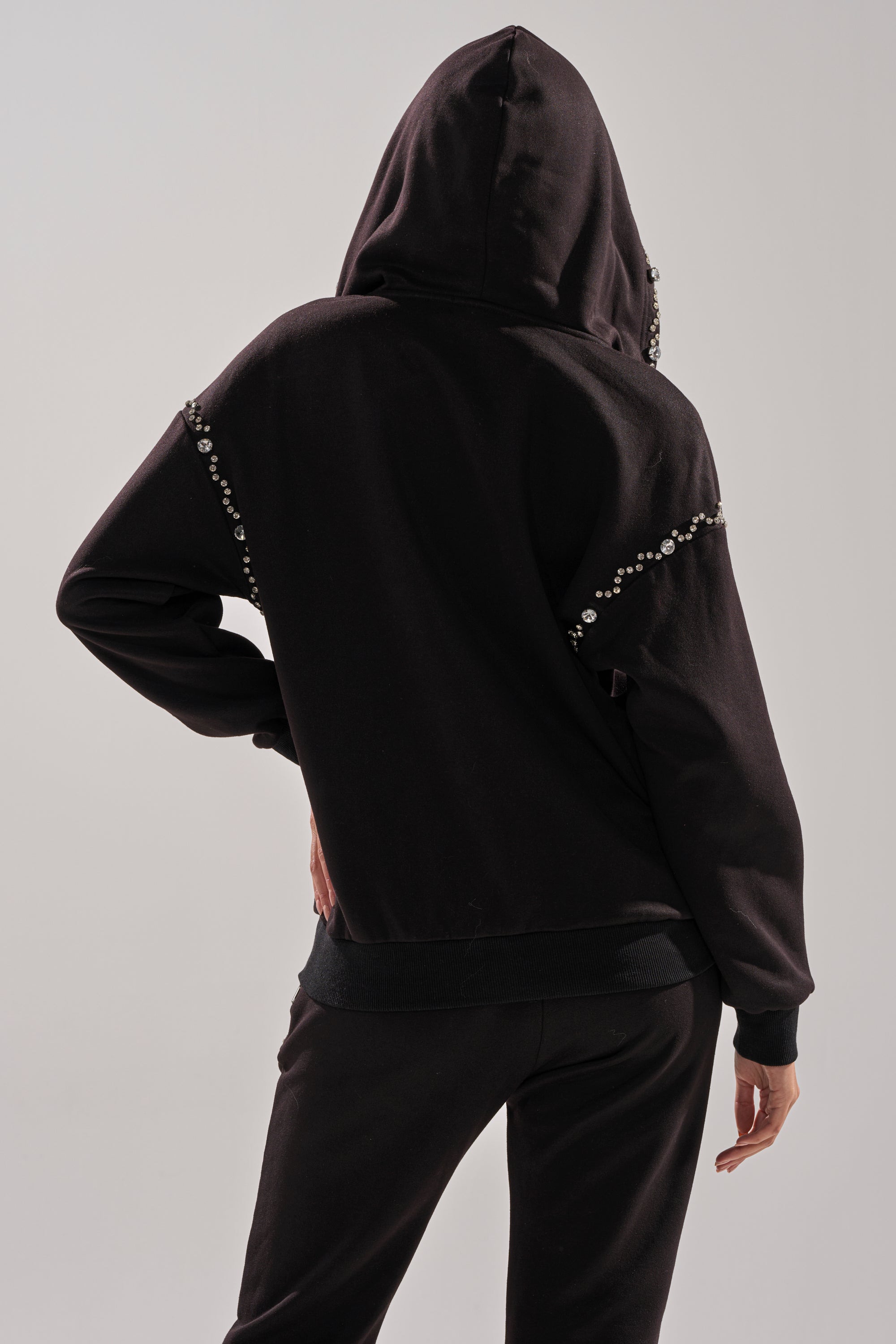 A person wearing the COZY HOLIDAY RHINESTONE ZIP UP SWEATSHIRT IN BLACK, featuring rhinestone embellishments on the sleeves, stands with one hand on their hip and their back to the camera.