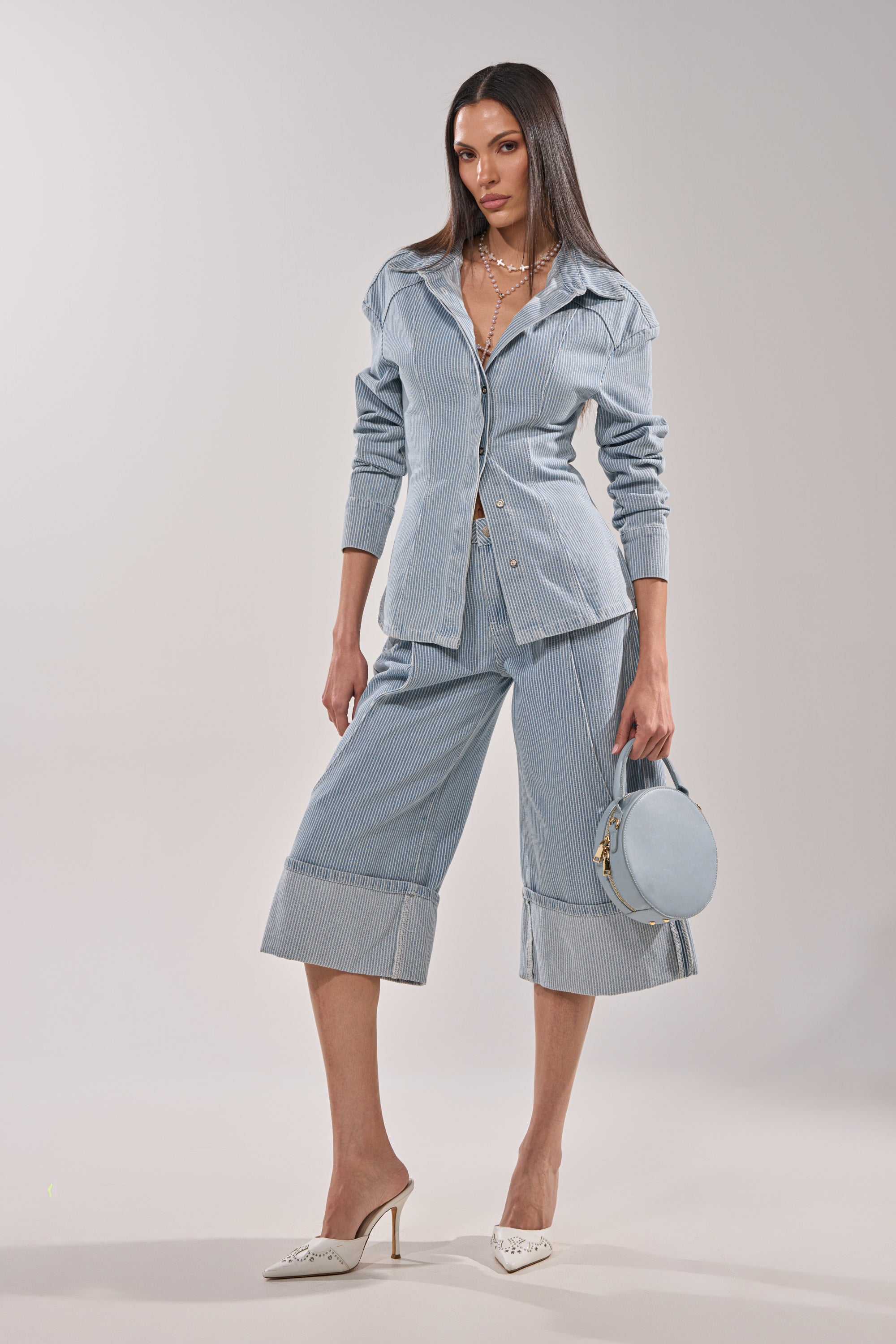 A woman models the FEEL FREE STRIPE GAUCHO set—a light blue pinstriped jacket and matching wide-leg cropped pants. She accessorizes with a pastel blue round handbag, white heels, and layered necklaces for a vintage-inspired style. 0