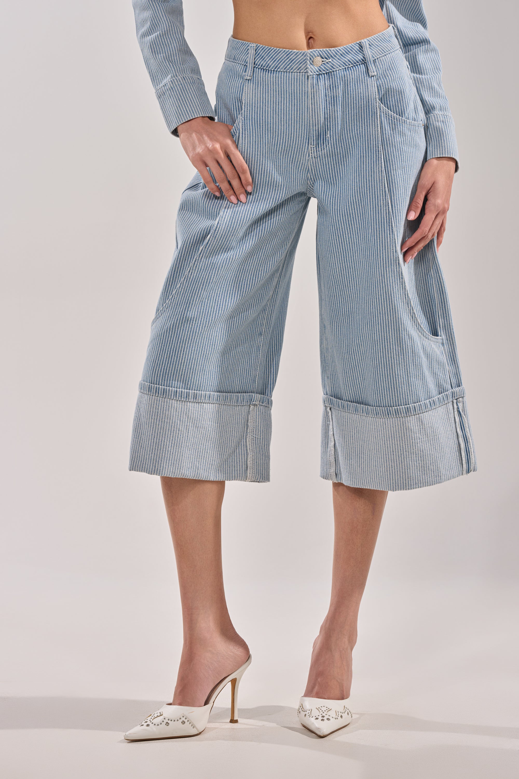 Wearing the FEEL FREE STRIPE GAUCHO—blue and white striped wide-leg cropped pants—a person sports a matching long-sleeve top and white high-heeled mules, posing against a plain background with their face not visible. 1