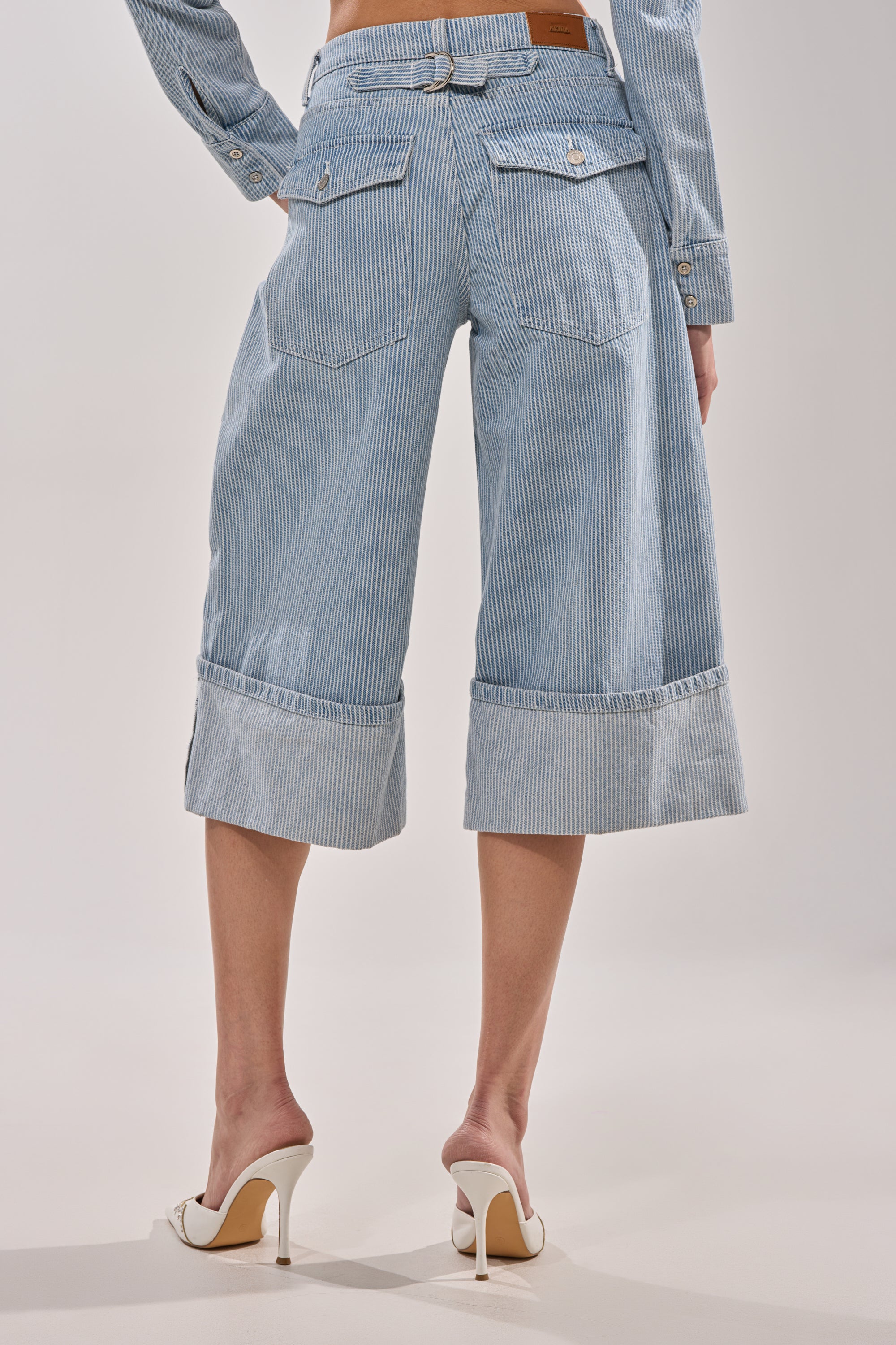 Someone wears the FEEL FREE STRIPE GAUCHO: blue-and-white pinstriped, mid-rise, wide-leg cropped pants with large back pockets and cuffs, styled with white high heels, photographed from behind against a plain backdrop. 5