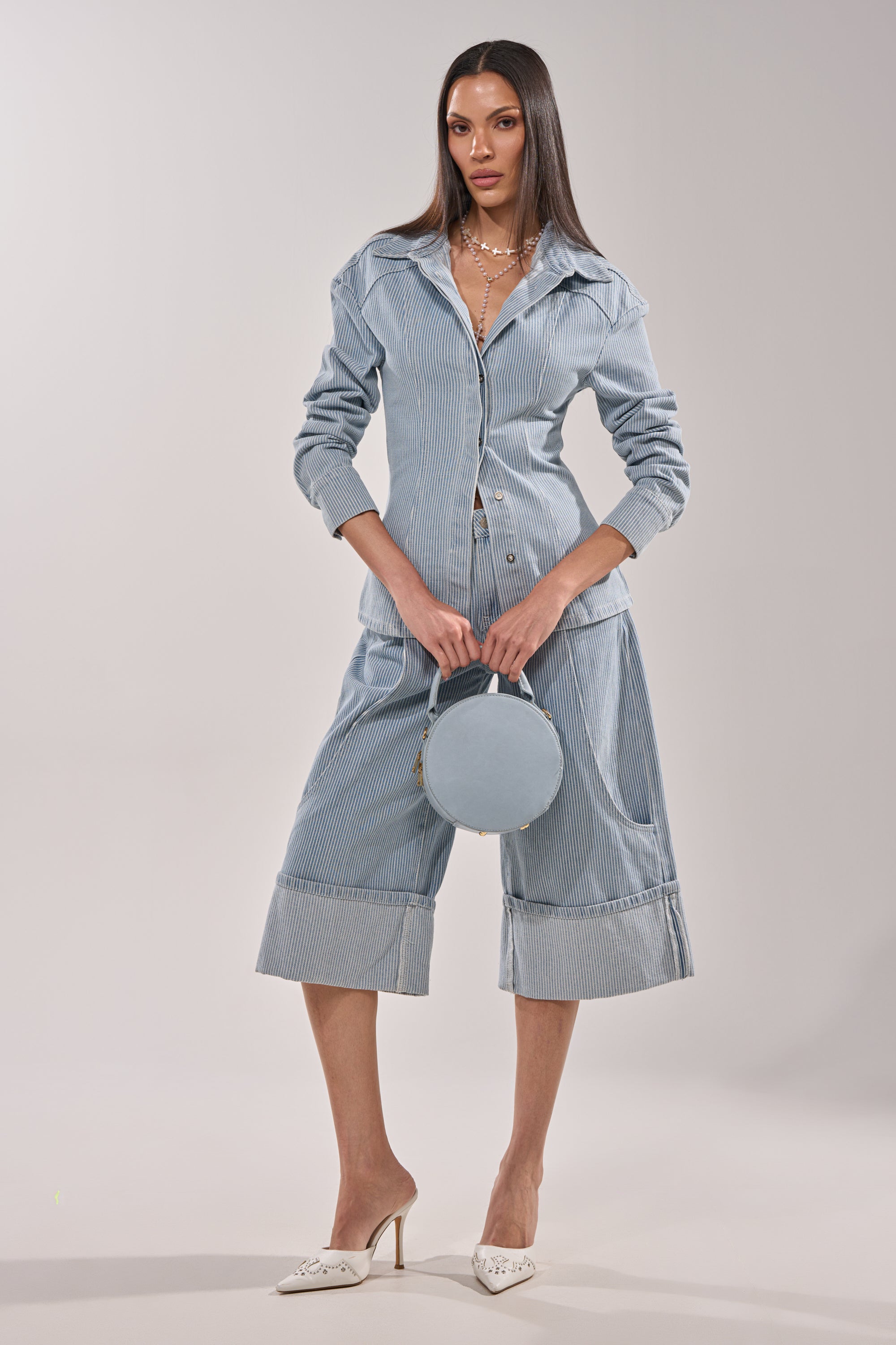 A woman stands confidently in the FEEL FREE STRIPE GAUCHO—vintage-inspired light blue pinstripe denim bermuda shorts paired with a matching button-up jacket, styled with a round blue handbag, white pointed heels, and layered necklaces. 6