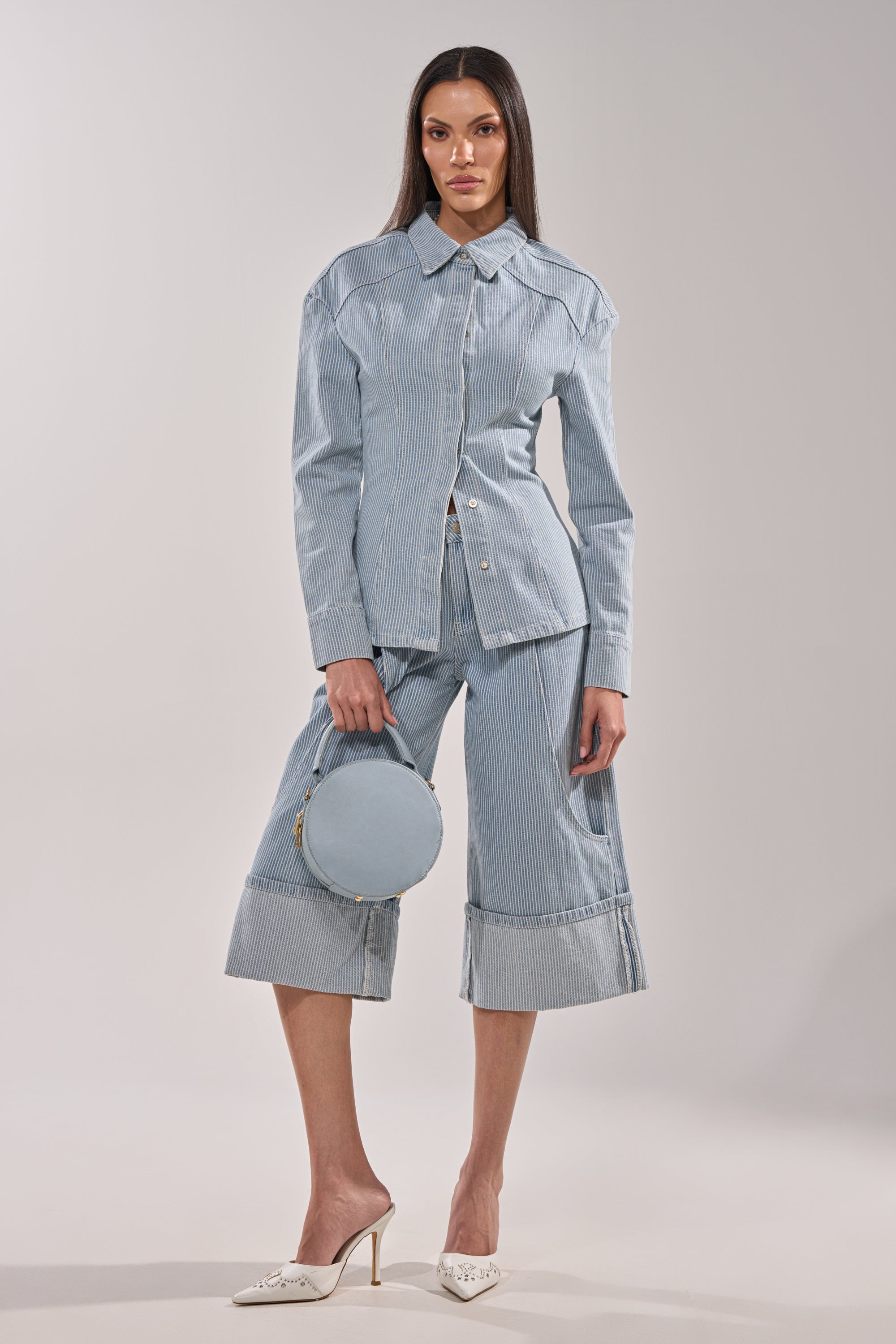 A woman wears the FEEL FREE STRIPE BUTTON DOWN, a light blue pinstripe blouse, with cropped matching wide-leg pants. She holds a round blue handbag and wears white pointed-toe heels against a plain, light-colored background. 9