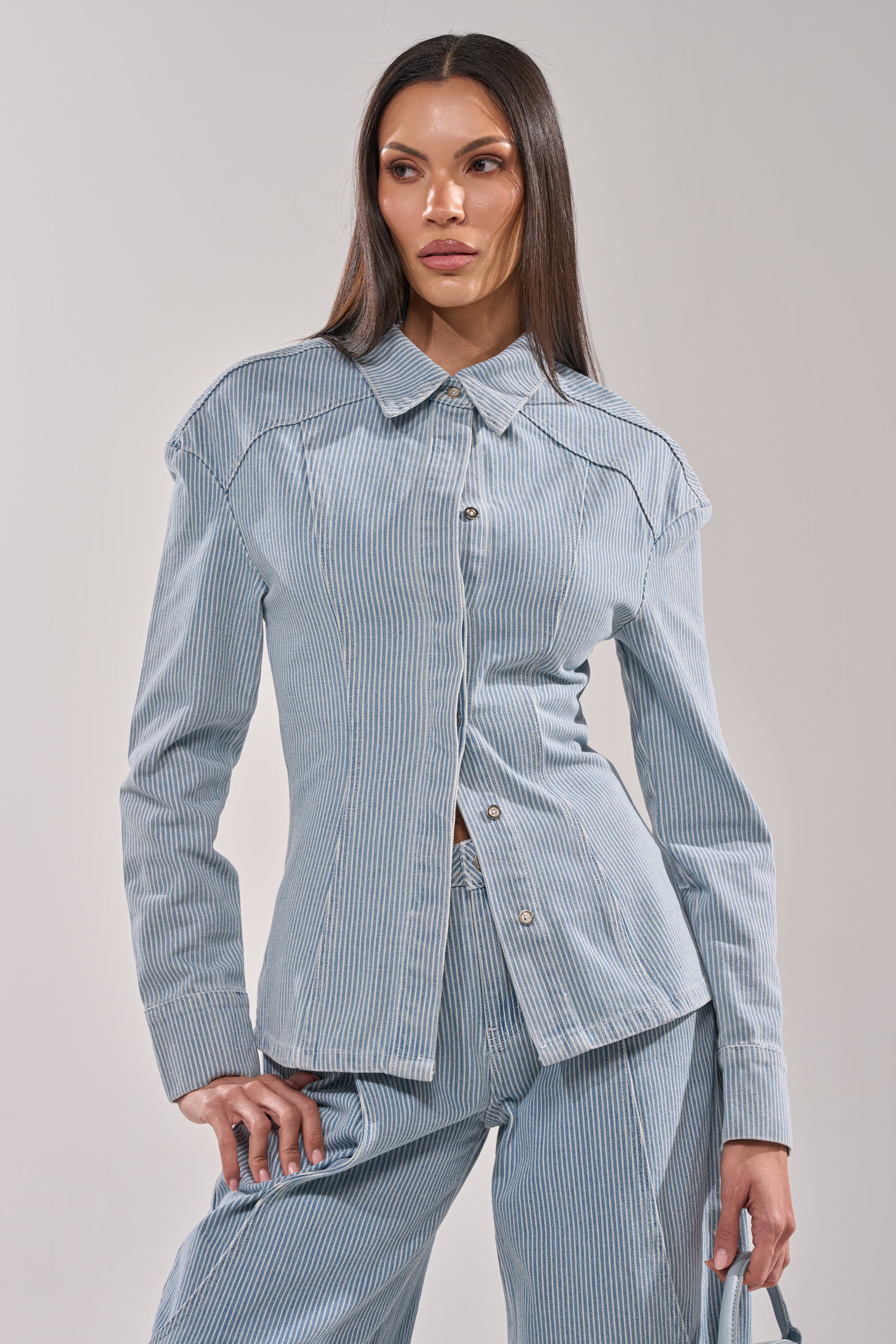 A woman with long straight hair poses against a plain background, wearing the FEEL FREE STRIPE BUTTON DOWN—a light blue and white pinstriped utility blouse—with matching pants, one hand on her hip and the other holding a bag. 1