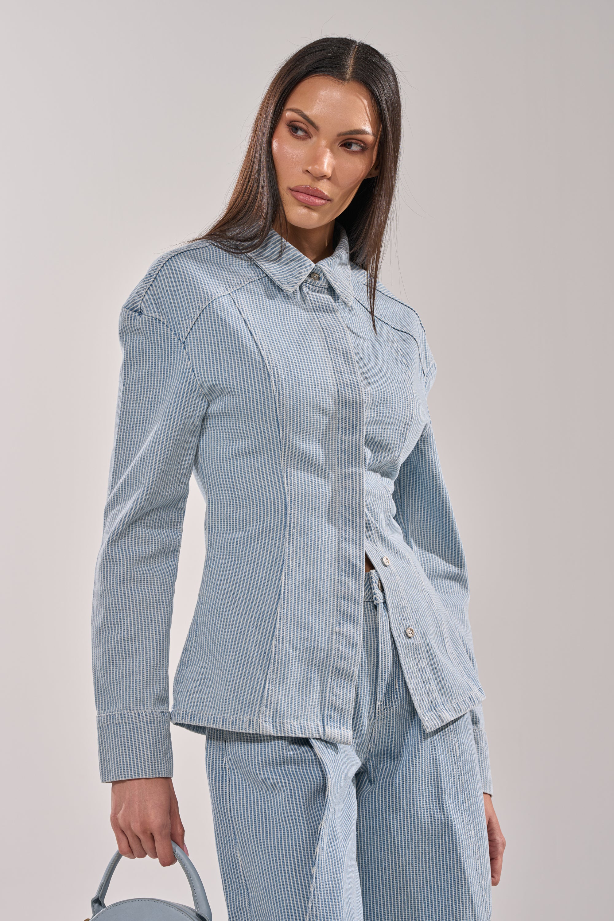 A woman with long straight hair wears the FEEL FREE STRIPE BUTTON DOWN, a light blue striped denim blouse, with matching pants and a small light blue handbag against a plain background. 3