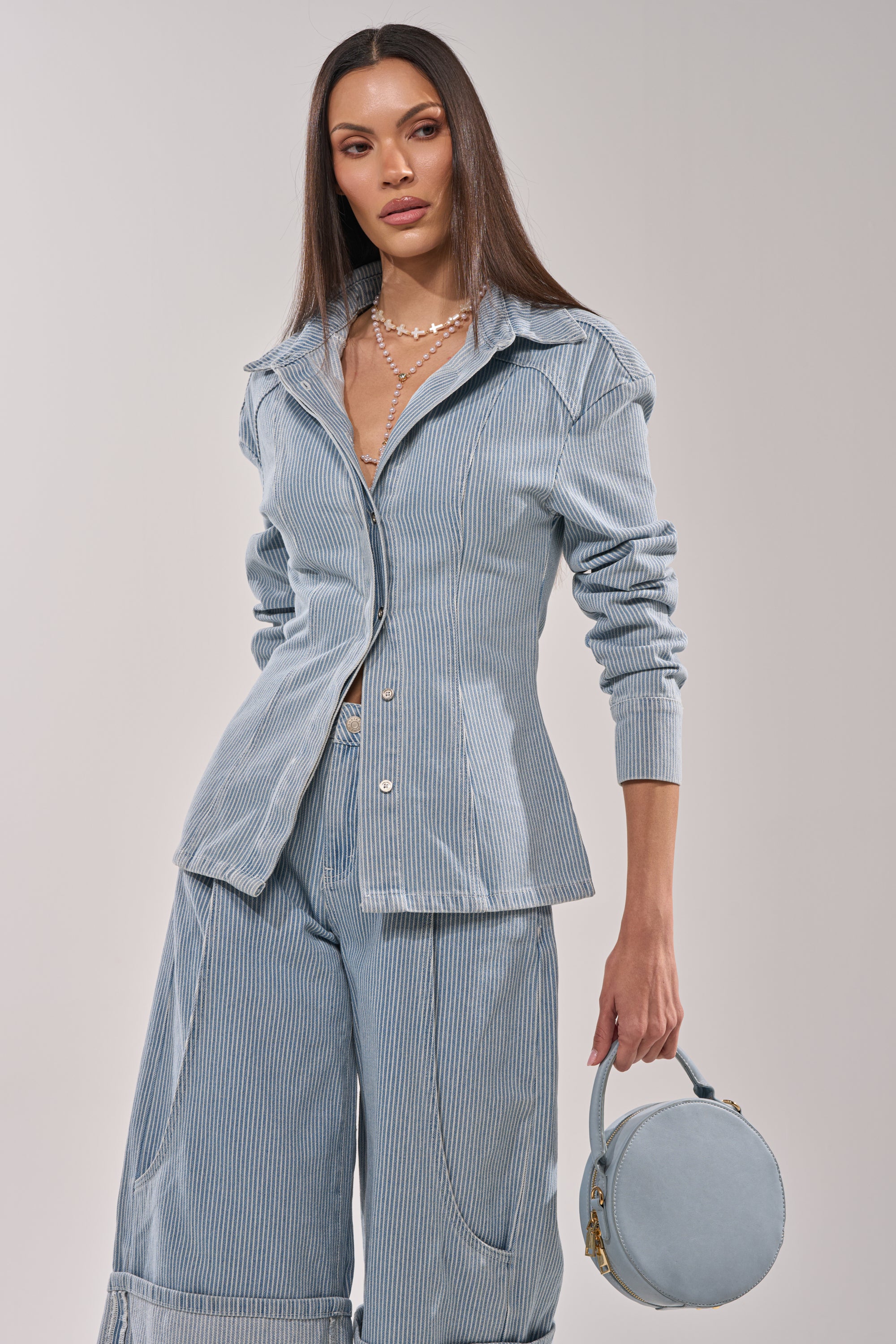 A woman wears the FEEL FREE STRIPE BUTTON DOWN—a light blue pinstriped blouse—styled with wide-leg pants and a matching round handbag. She accessorizes with layered pearl necklaces and poses against a plain light background. 4