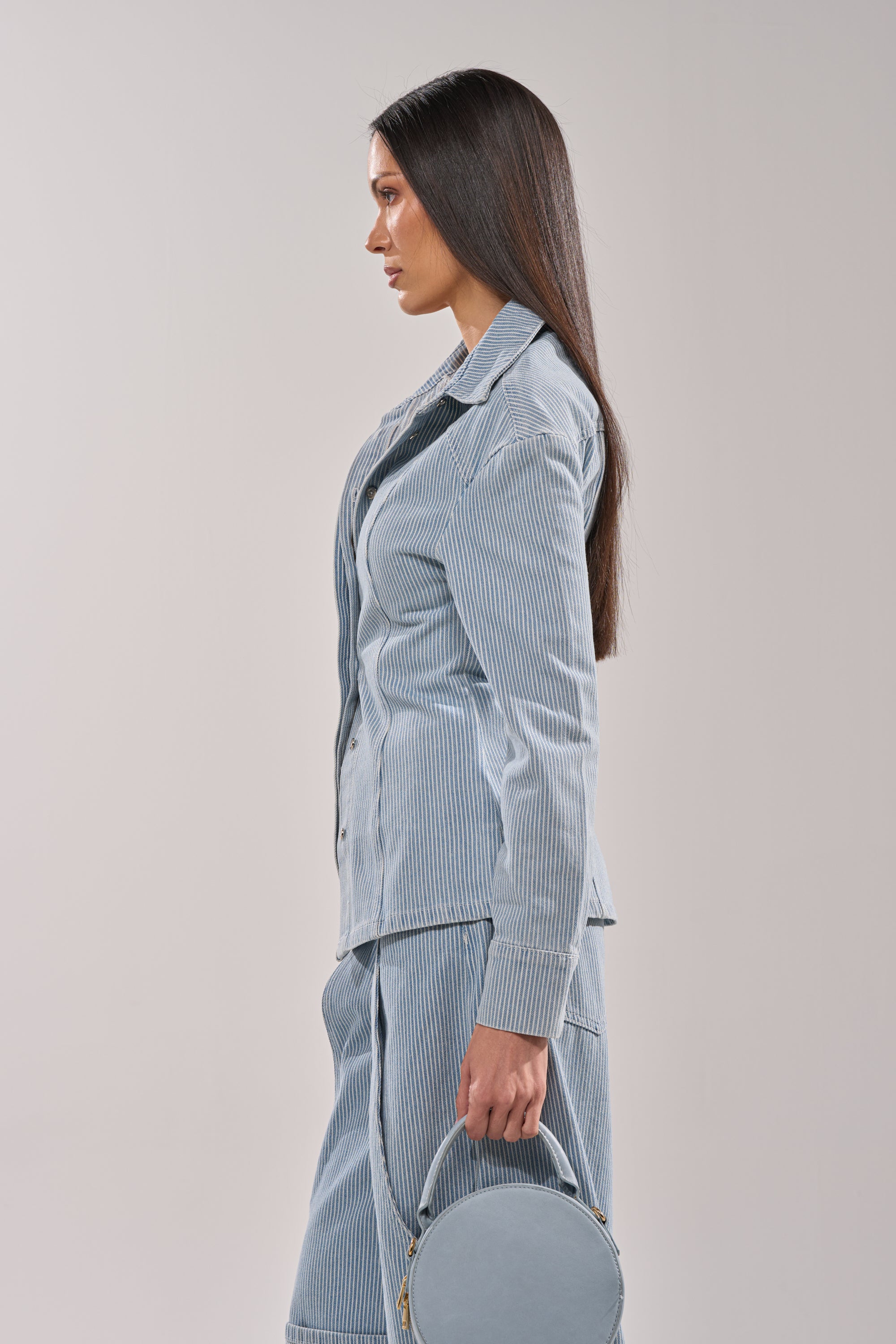 Woman with long straight hair stands in profile, wearing the FEEL FREE STRIPE BUTTON DOWN in light blue with matching pants and holding a round light blue handbag against a plain, light background. 5