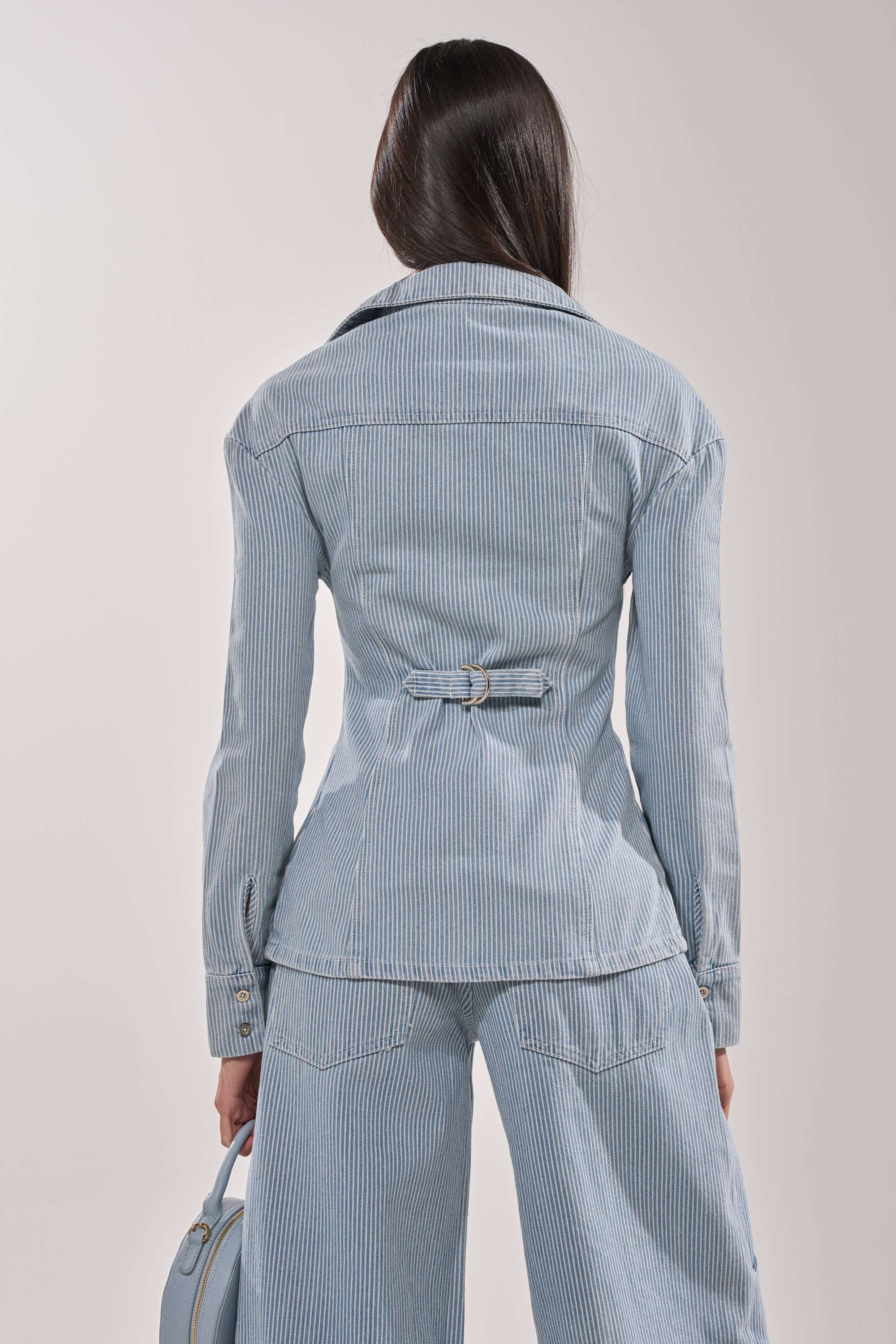 Woman with long dark hair, seen from behind, wearing the FEEL FREE STRIPE BUTTON DOWN in light blue stripes with matching wide-leg pants and holding a small light blue bag in her left hand. 6