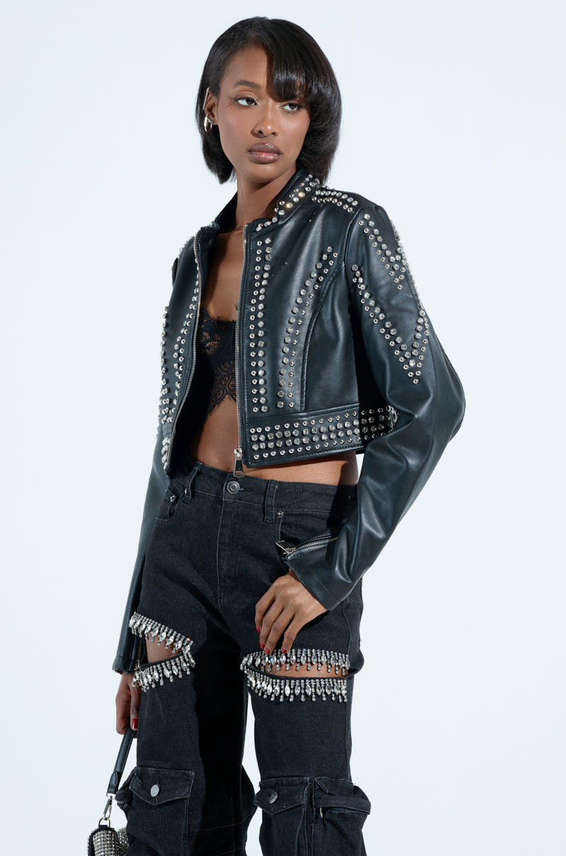 Wearing the STEPPING OUT EMBELLISHED MOTO studded faux leather jacket over a lacy top, paired with black pants featuring tear-away sections and hanging beaded fringe, they hold a small purse against a plain background.
