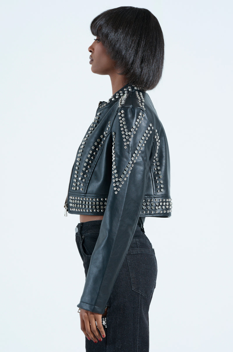 With chin-length hair, a person sports the STEPPING OUT EMBELLISHED MOTO jacket, featuring intricate spike patterns and a metallic zip closure, paired with dark jeans. Posed in profile against a plain backdrop, this black ensemble showcases bold fashion statements.