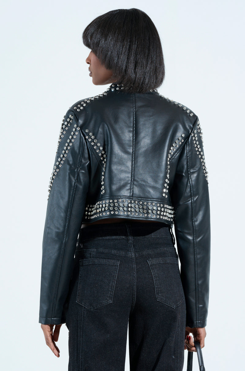 A person with short straight hair is seen from behind wearing the STEPPING OUT EMBELLISHED MOTO jacket, featuring a metallic zip closure, paired with black jeans. They hold a small handbag against a plain white background.