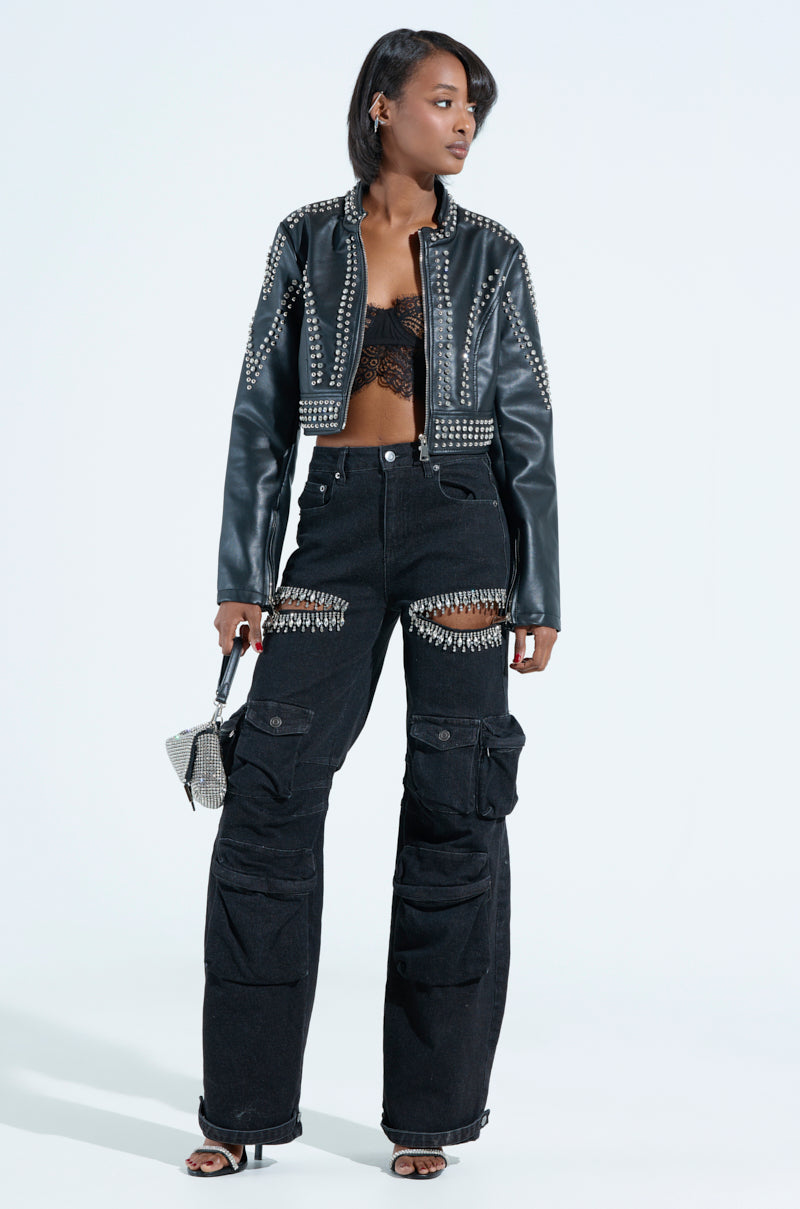 A woman confidently models the STEPPING OUT EMBELLISHED MOTO jacket with metallic zippers and black cargo pants adorned with beads. She accessorizes with a sparkly handbag, wears heels, and showcases a lace top under the faux leather jacket against a plain white backdrop.