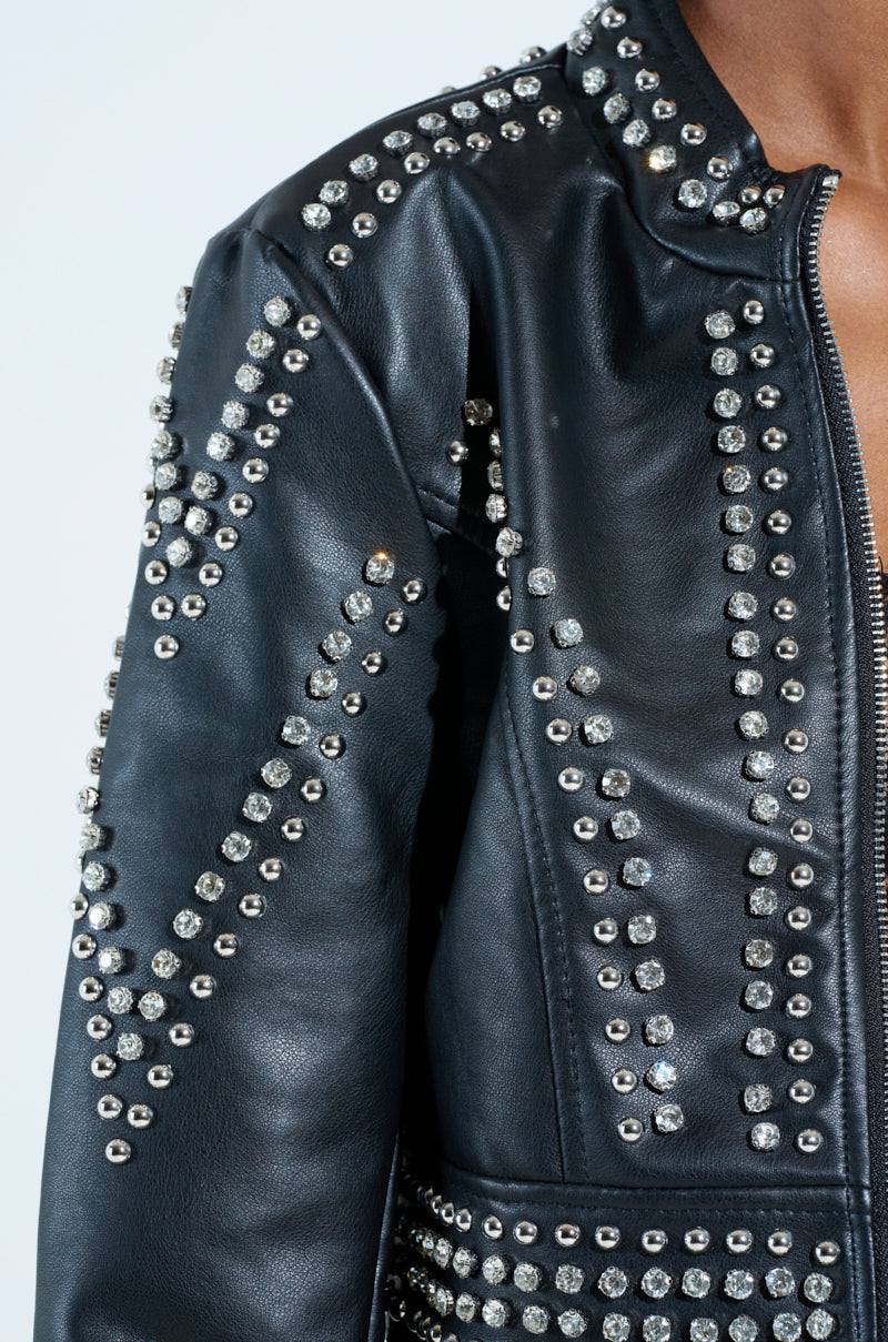 The STEPPING OUT EMBELLISHED MOTO jacket in black faux leather showcases silver studs in geometric patterns, complemented by a metallic zip closure, offering a bold and edgy look.