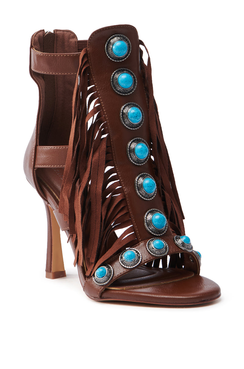 The AZALEA WANG SUMAC BROWN WESTERN SANDAL is a brown, gladiator-style high-heeled open-toe faux leather shoe with fringe design, back zipper, slim heel, and embellished with large turquoise stones in circular silver settings.