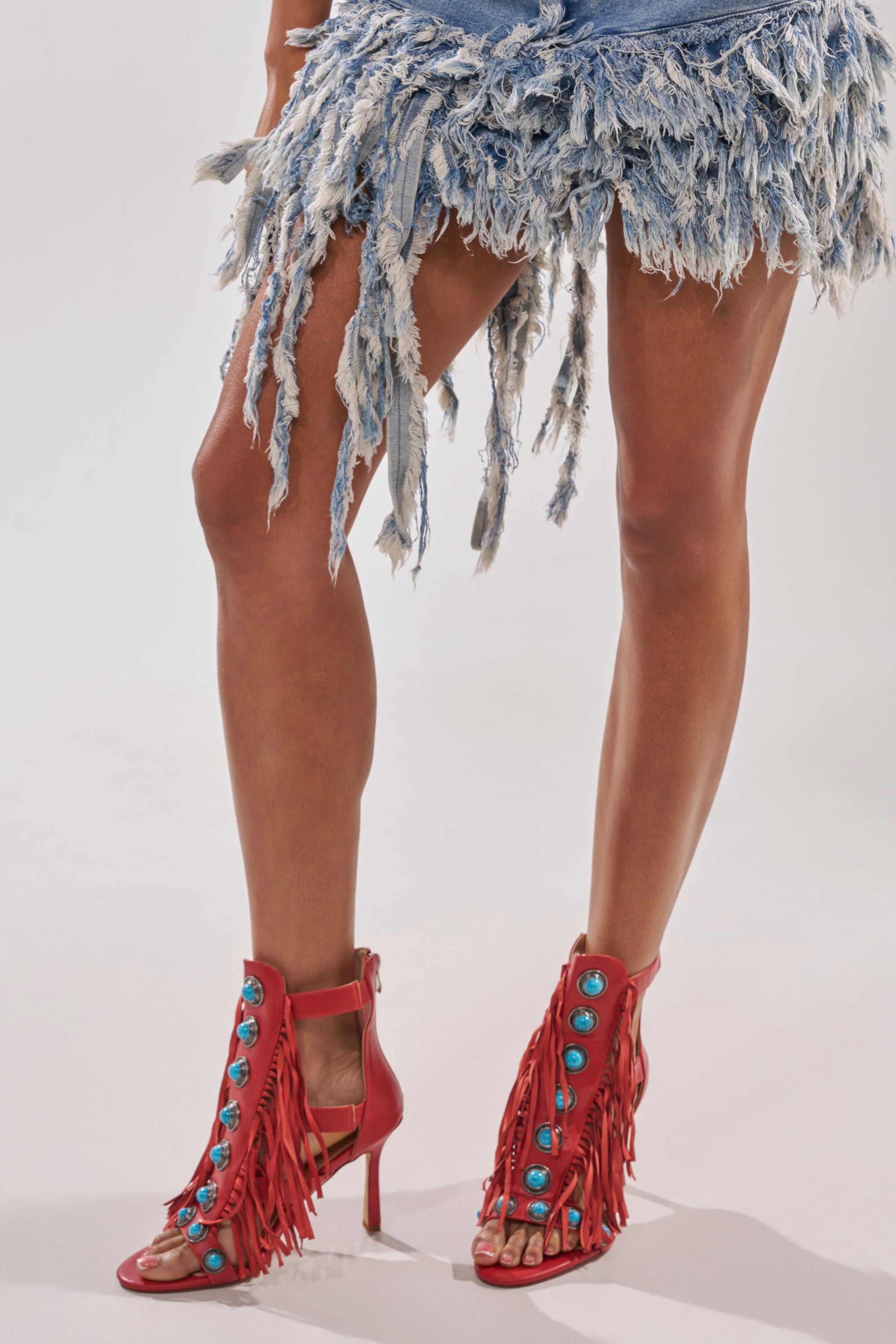 A person poses against a plain background wearing a distressed, fringed denim skirt and the AZALEA WANG SUMAC RED FRINGE WESTERN STILETTO SANDAL, featuring bold turquoise stones and statement fringe details. 0