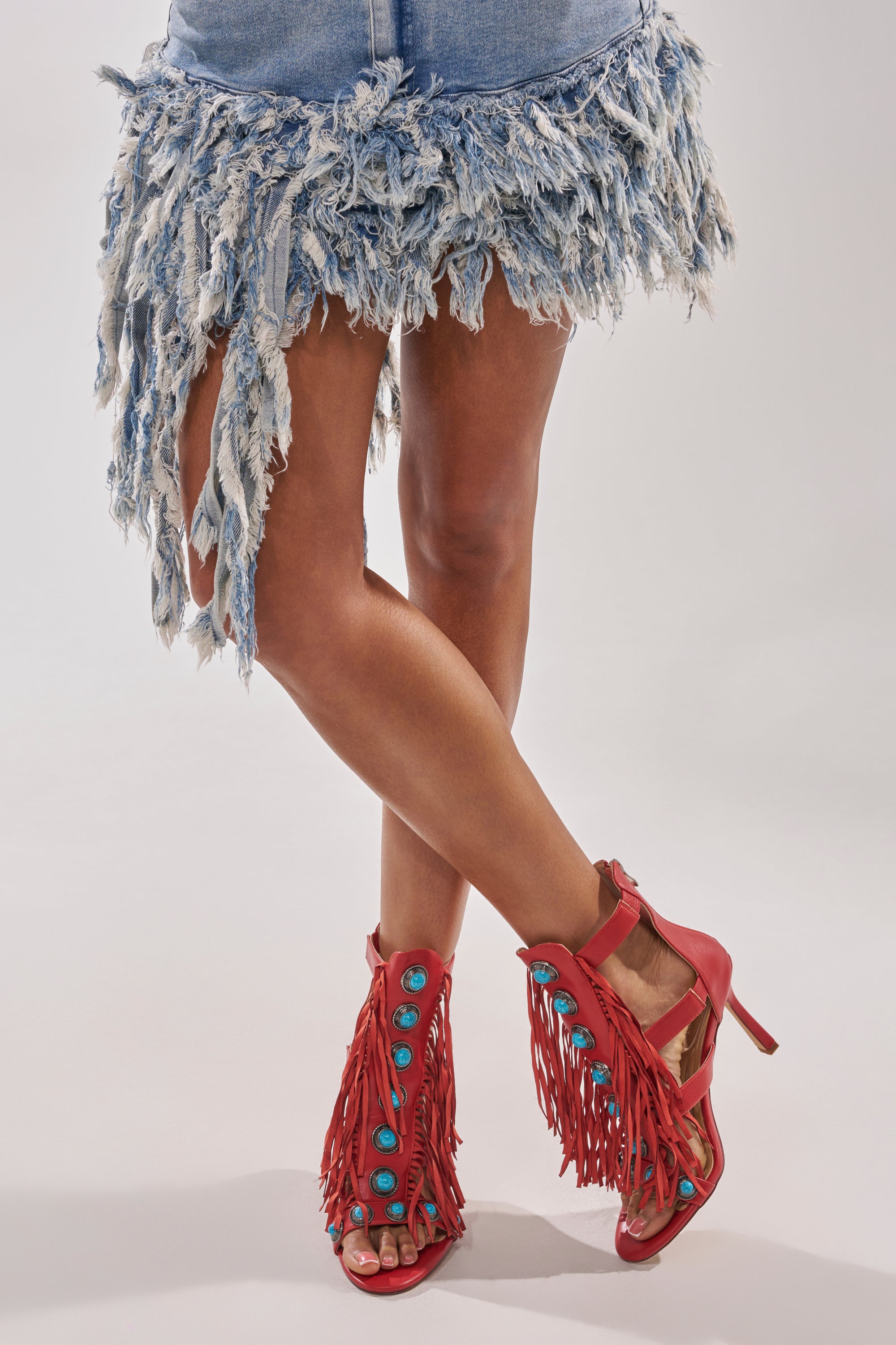 A person in a frayed denim skirt stands with legs crossed, showcasing the AZALEA WANG SUMAC RED FRINGE WESTERN STILETTO SANDAL—bold red heels featuring fringe and turquoise stones—against a plain background. 1