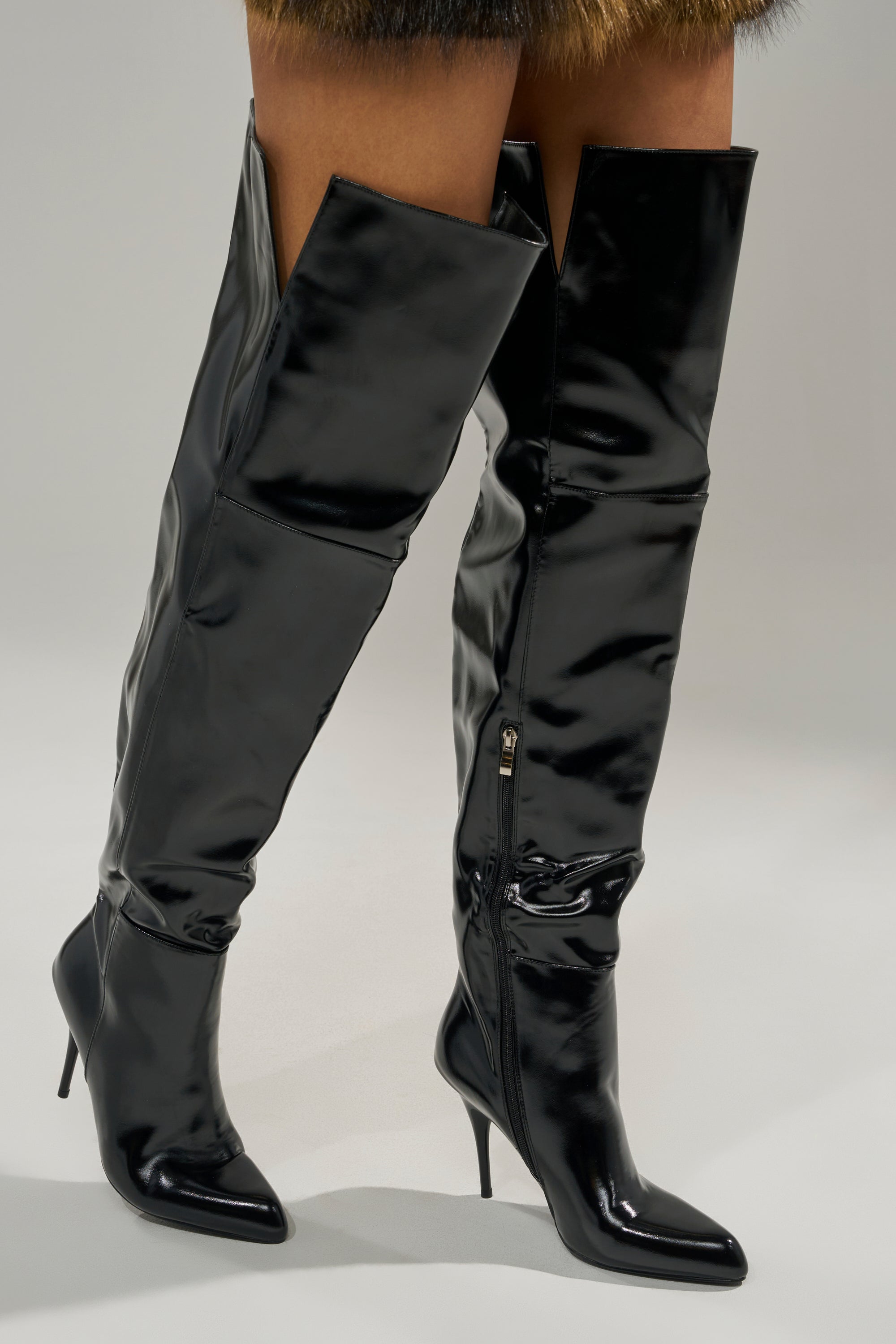 A person wearing the AZALEA WANG SUNSHINE THIGH HIGH BLACK PU STILETTO BOOT with pointed toes and stiletto heels stands on a light gray surface.