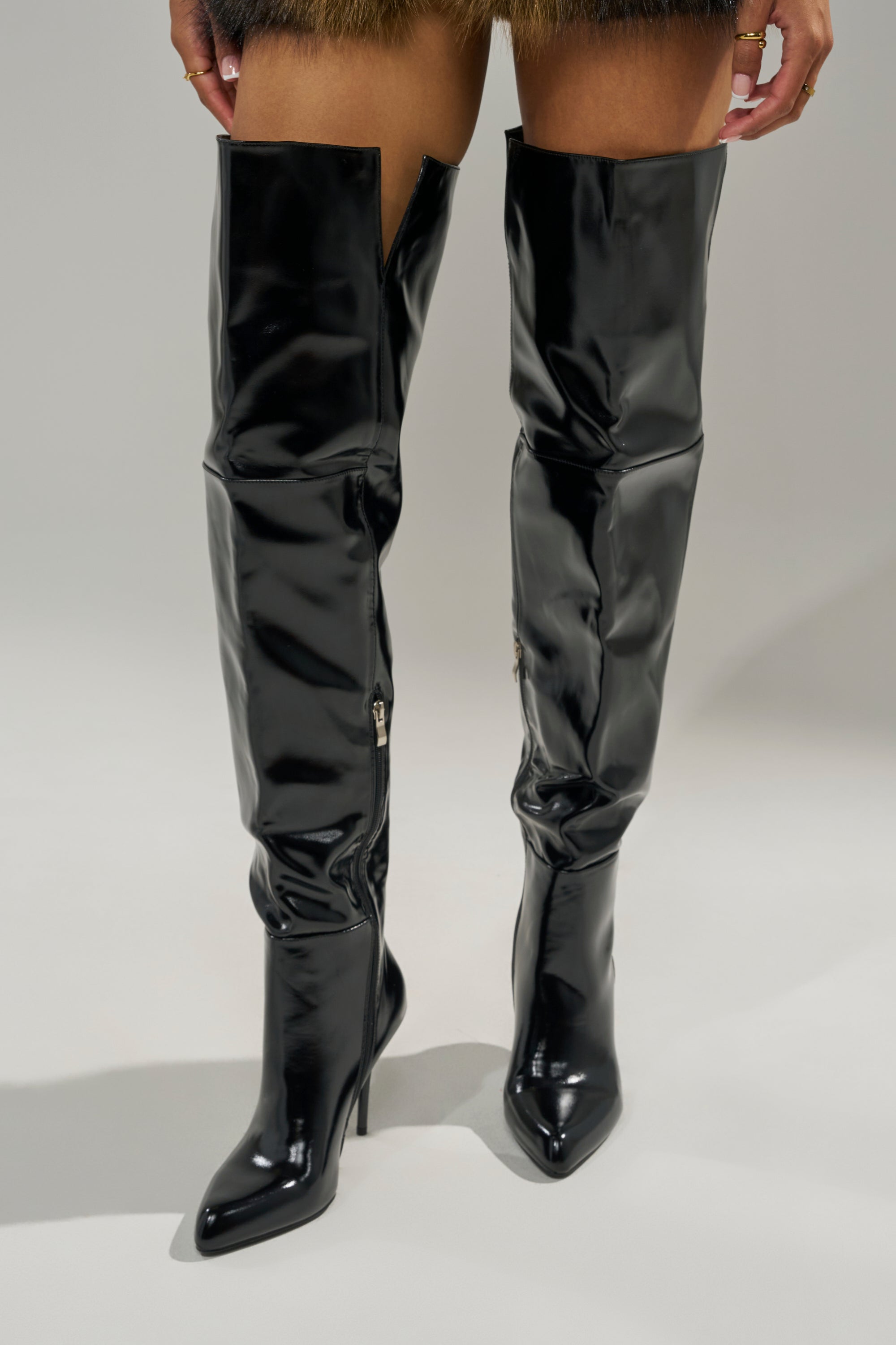A person is standing on a light surface wearing AZALEA WANG SUNSHINE THIGH HIGH BLACK PU STILETTO BOOTS, with only the lower legs and the shiny, over-the-knee boots visible.