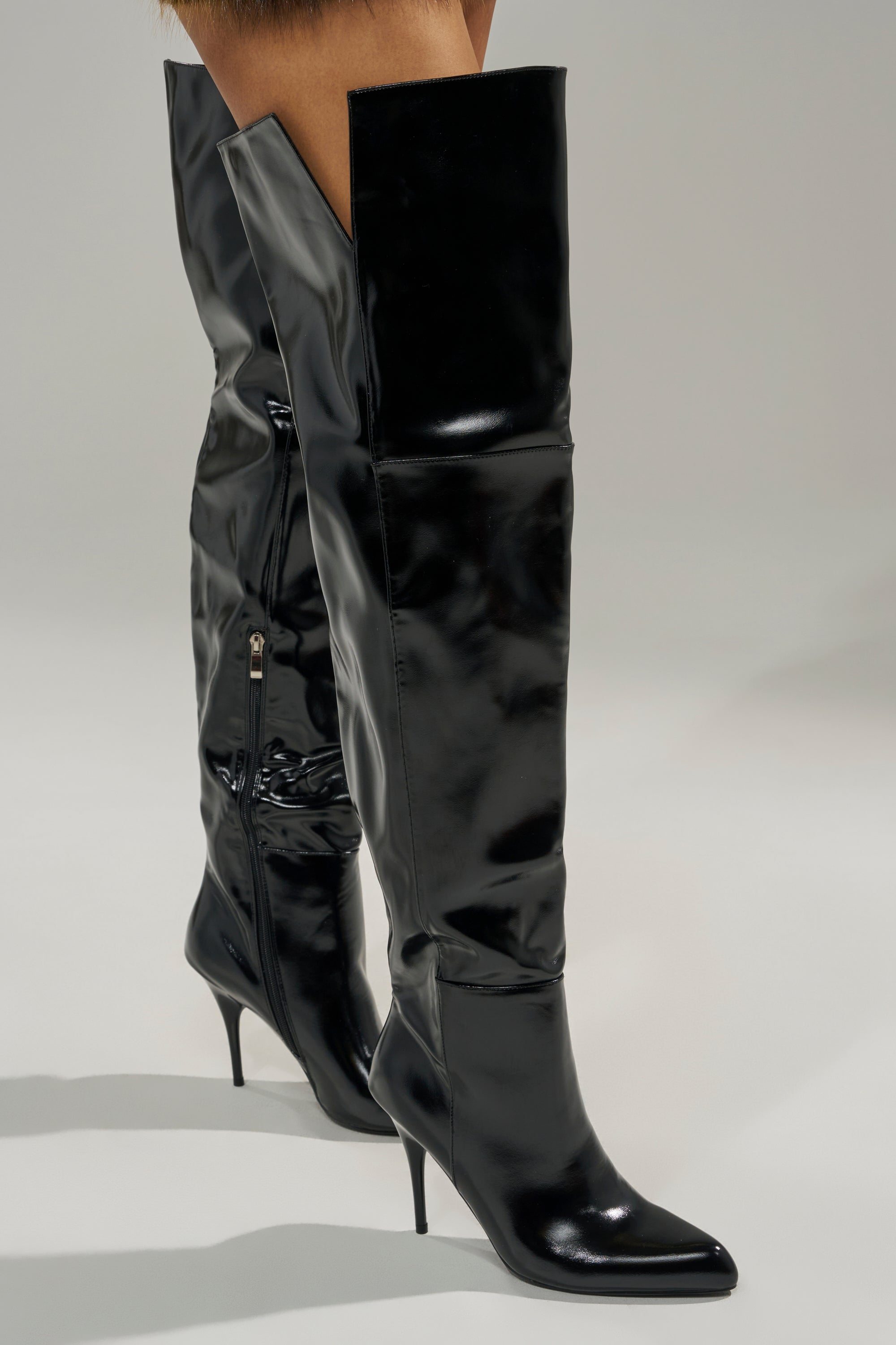 A person stands on a light gray surface wearing the AZALEA WANG SUNSHINE THIGH HIGH BLACK PU STILETTO BOOT, featuring shiny black PU, pointed toes, high stiletto heels, and side zippers.