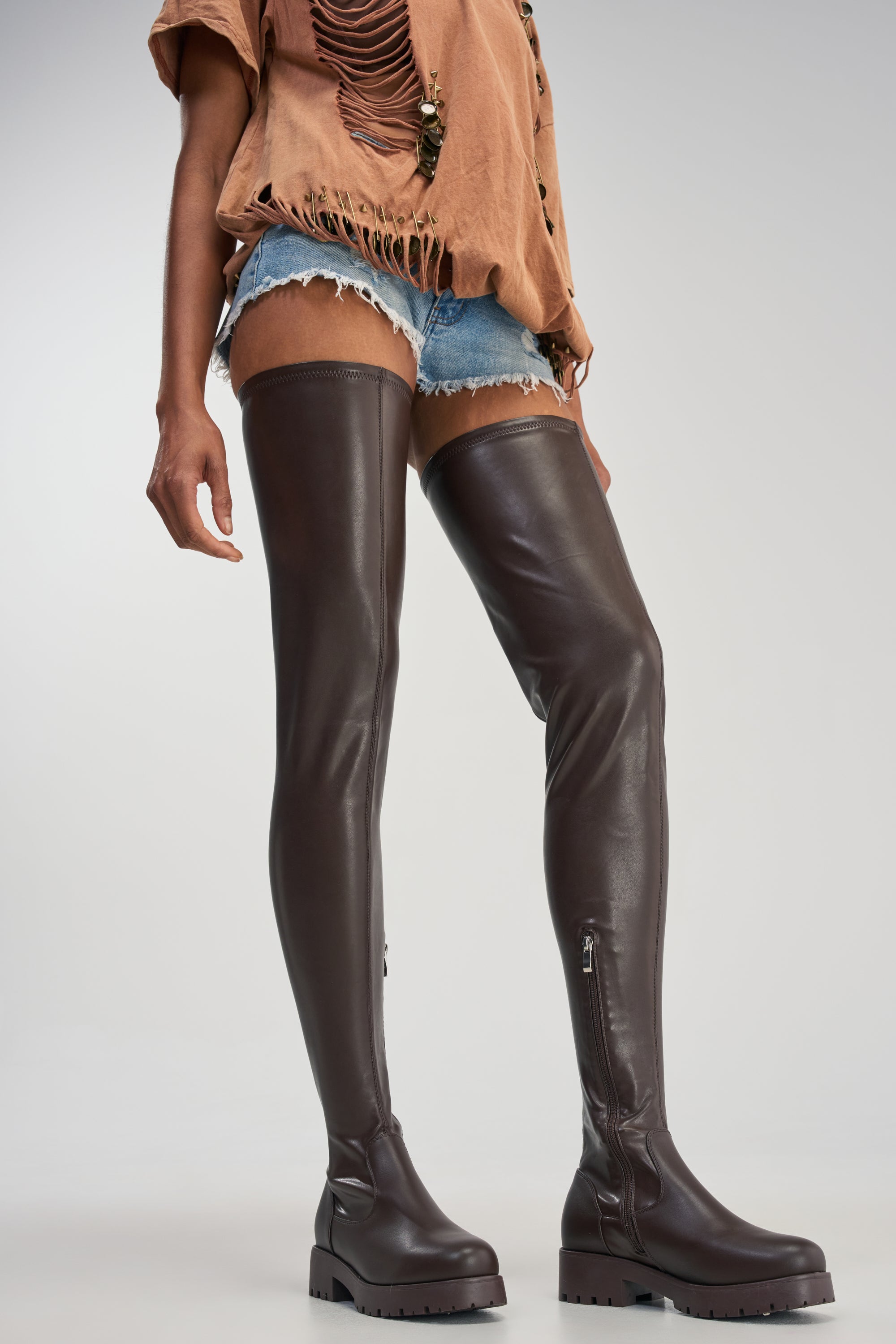 A person wearing a distressed tan t-shirt, denim shorts, and the AZALEA WANG SURGICAL NEW THIGH HIGH STRETCH PU FLAT BOOT in chocolate brown with a platform lug sole, standing against a plain background.