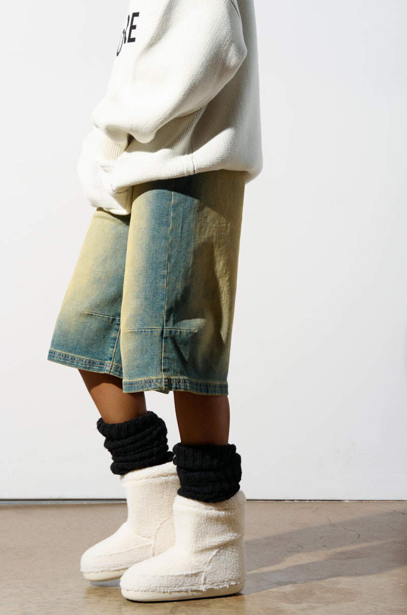 A person wearing an oversized white sweater pairs it with faded denim shorts, thick black leg warmers, and the cozy AZALEA WANG SWEETING WHITE SHERPA BOOTIE. The backdrop is a plain, light-colored wall and floor, emphasizing the textured flatform soles visible beneath the fluffy slippers.