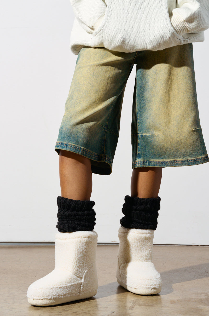 A person wearing a white hoodie and knee-length denim shorts stands indoors, their look completed with black knitted leg warmers and the AZALEA WANG SWEETING WHITE SHERPA BOOTIE. The background is a plain white wall and a light-colored floor.