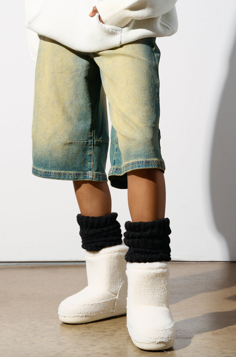 A person wearing faded denim shorts, a white sweater, black leg warmers, and AZALEA WANG SWEETING WHITE SHERPA BOOTIES with an ankle-high shaft is standing on a beige floor. The focus is on their lower half, creating a casual and comfortable look.