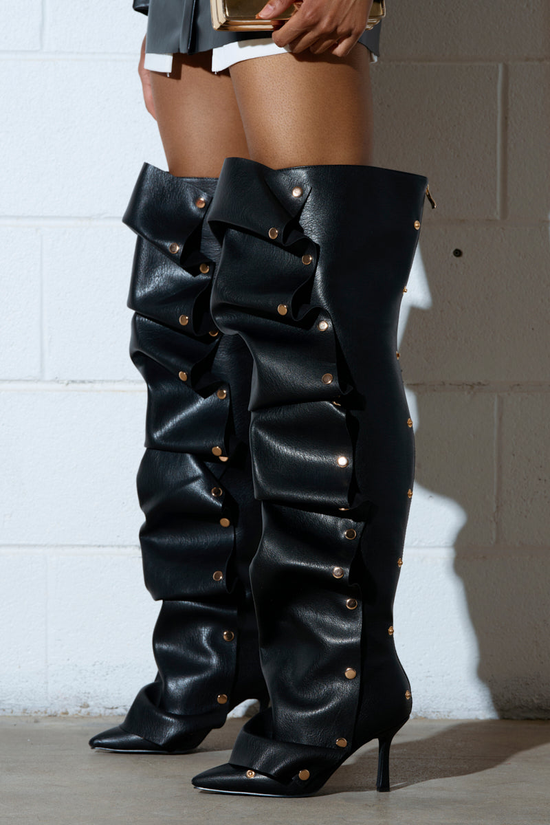 The AZALEA WANG SWELCIE BLACK PU GOLD HARDWARE STILETTO BOOTS, with ruffled detailing and gold buttons, are strikingly extravagant against a white brick wall background.