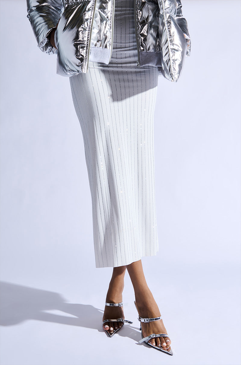 A person is wearing a metallic silver jacket, a white skirt with vertical pinstripes, and AZALEA WANG SWEN SILVER STILETTO SANDALS. The pointed open-toe design complements their look as the image captures their lower half against a light background.