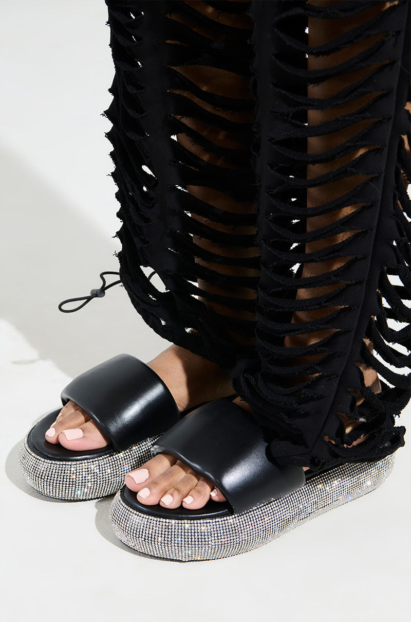 Wearing AZALEA WANG SWITZERLAND BLACK RHINESTONE SANDALS with thick flatform soles, a person enhances a chic black shredded-length dress, revealing painted toenails. Made from vegan leather, the sandals complement the minimalistic background perfectly.