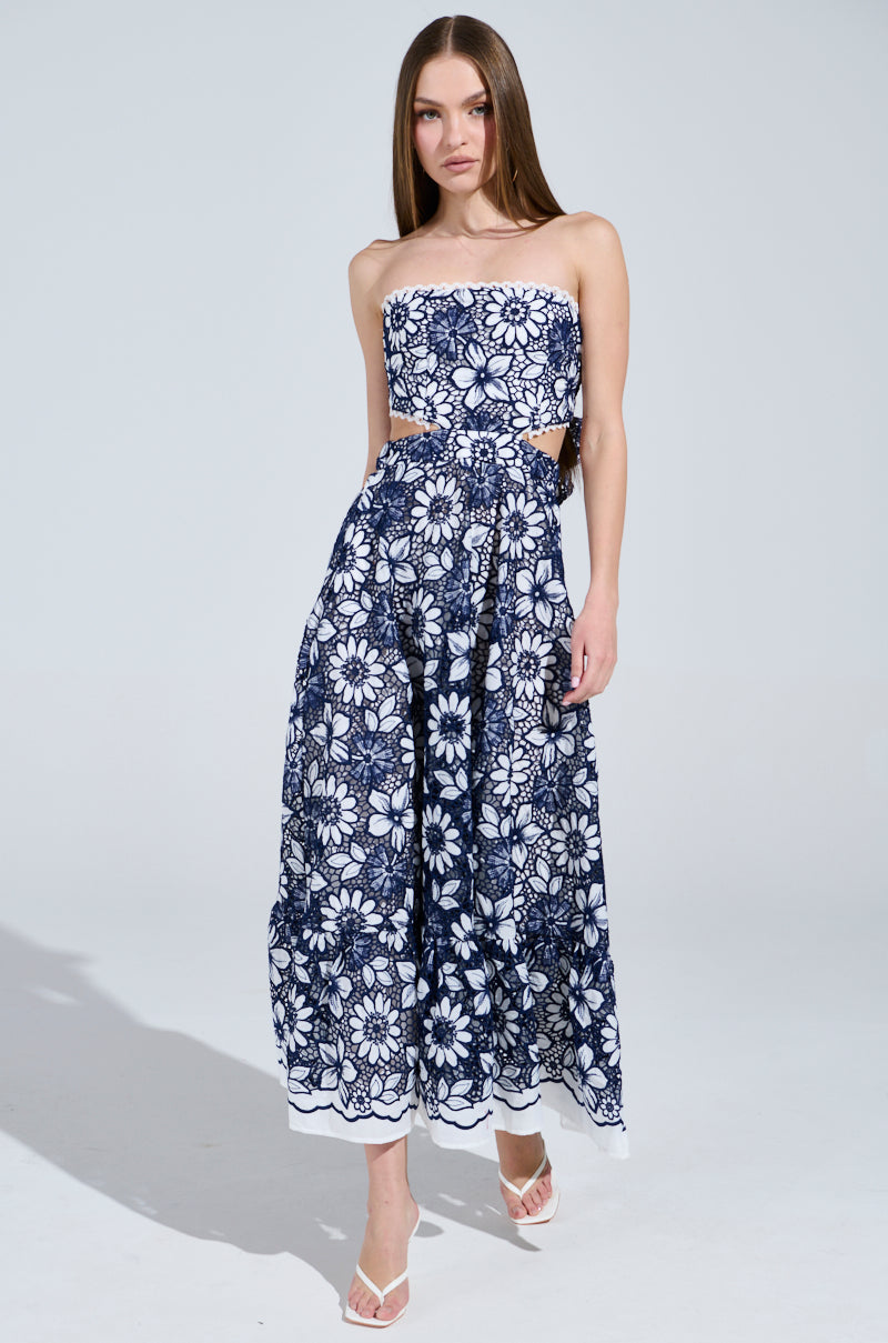 A woman poses against a plain background in the HERE TO CELEBRATE FLORAL MIDI DRESS, featuring a strapless neckline, ankle-length blue and white floral design with side cutouts and a delicate crochet pattern. Her look is complemented by stylish white sandals.