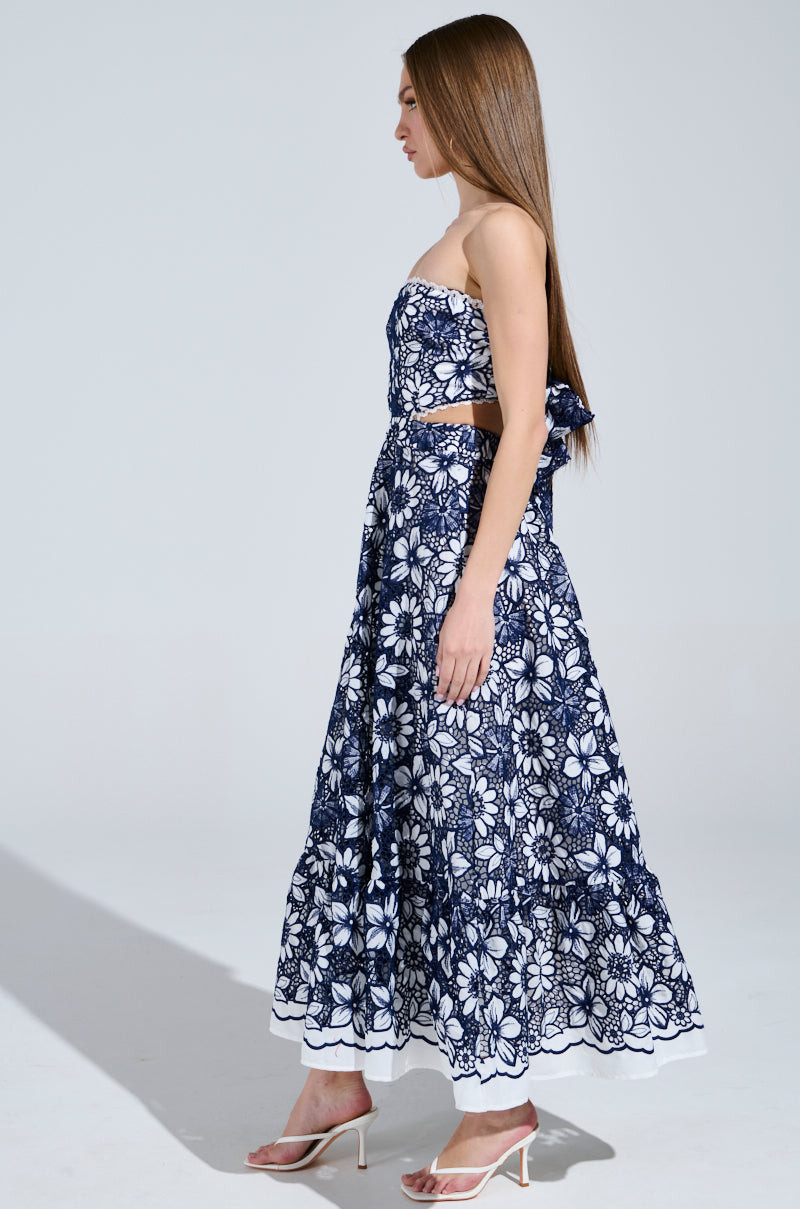 A woman in the HERE TO CELEBRATE FLORAL MIDI DRESS, featuring a strapless navy blue and white floral design, stands sideways looking forward. She wears white heels, has long brown hair, and casts a shadow on a plain background.