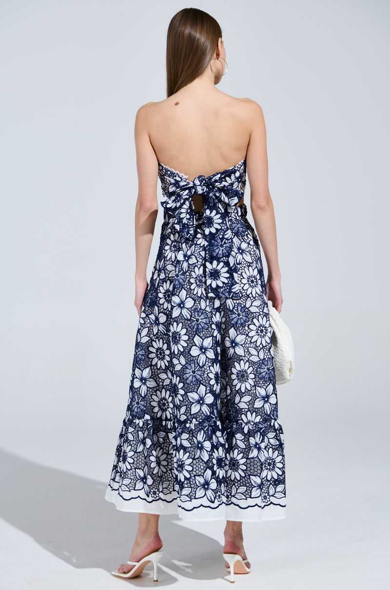 A woman with long brown hair showcases the HERE TO CELEBRATE FLORAL MIDI DRESS, featuring a strapless navy blue and white floral design with a delicate crochet pattern. She accessorizes with a white purse and wears white high-heeled sandals.