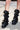 A person stands on a sidewalk wearing the AZALEA WANG SYBEL BUTTON FLAT BOOTIE—tall, slouchy black boots with platform lug soles, exaggerated folds, and metal rivets. Only the lower legs are visible.