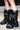 Close-up of a person wearing the AZALEA WANG SYBEL BUTTON FLAT BOOTIE, a black faux leather bootie with metallic snap buttons and a thick platform lug sole, standing on a sidewalk with an urban backdrop.