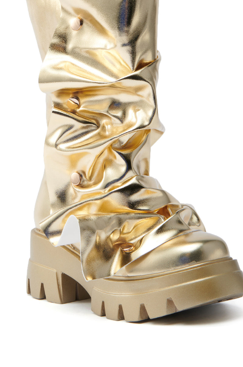 The AZALEA WANG SYBEL BUTTON GOLD FLAT BOOTIE is a close-up of a metallic bootie with a crumpled texture, platform lug sole, and glossy faux leather. It has decorative studs along its gathered sections.