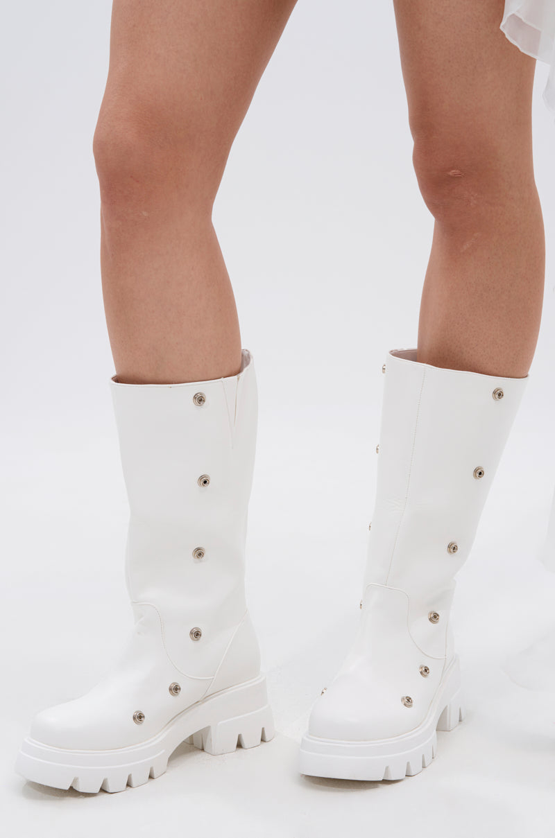 A person models the AZALEA WANG SYBEL WHITE SNAP DETAIL BOOTIE—knee-high, faux leather boots with chunky heels and decorative metal snaps—against a white background. Only their lower legs and the bold boots are visible.
