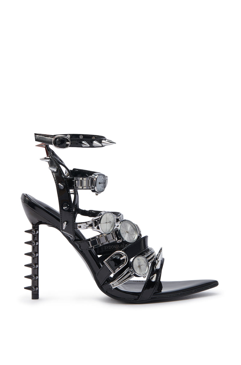 The AZALEA WANG SYDONDIA BLACK WATCH SHOE STILETTO SANDAL is a high-heeled faux patent leather sandal with adjustable ankle straps, silver buckles, pointed spikes, and metallic watch faces for an edgy industrial look.