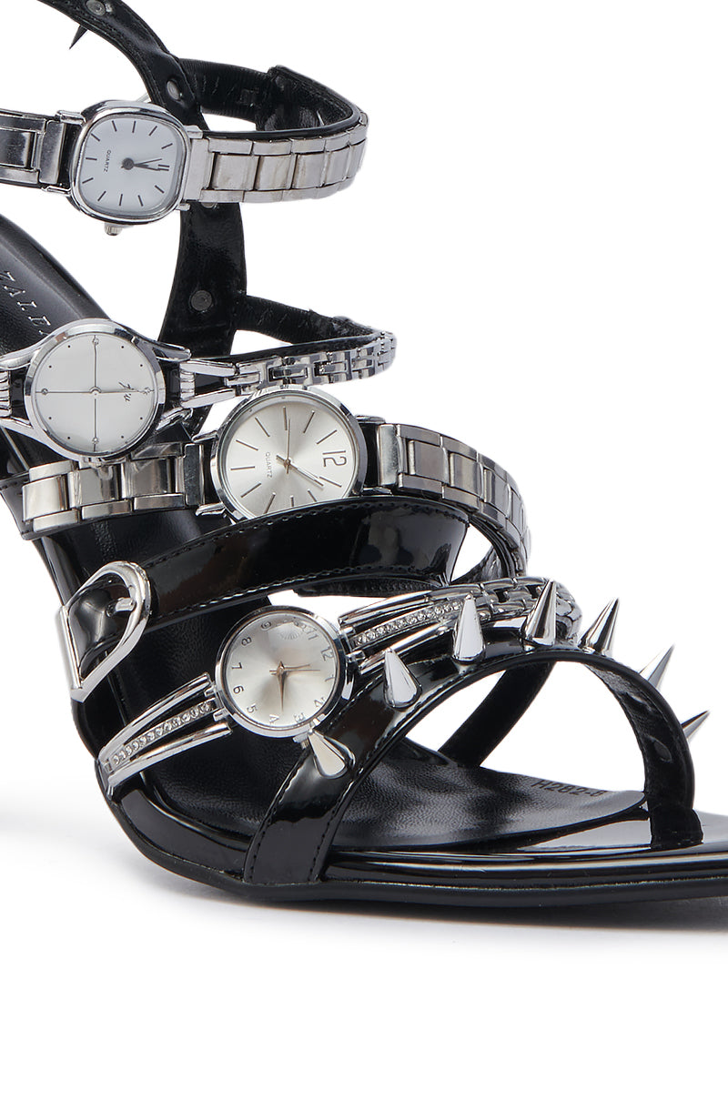 The AZALEA WANG SYDONDIA BLACK WATCH SHOE STILETTO SANDAL features black patent faux leather, multiple adjustable ankle straps, and edgy silver wristwatch and metal spike details for a bold, unconventional look.