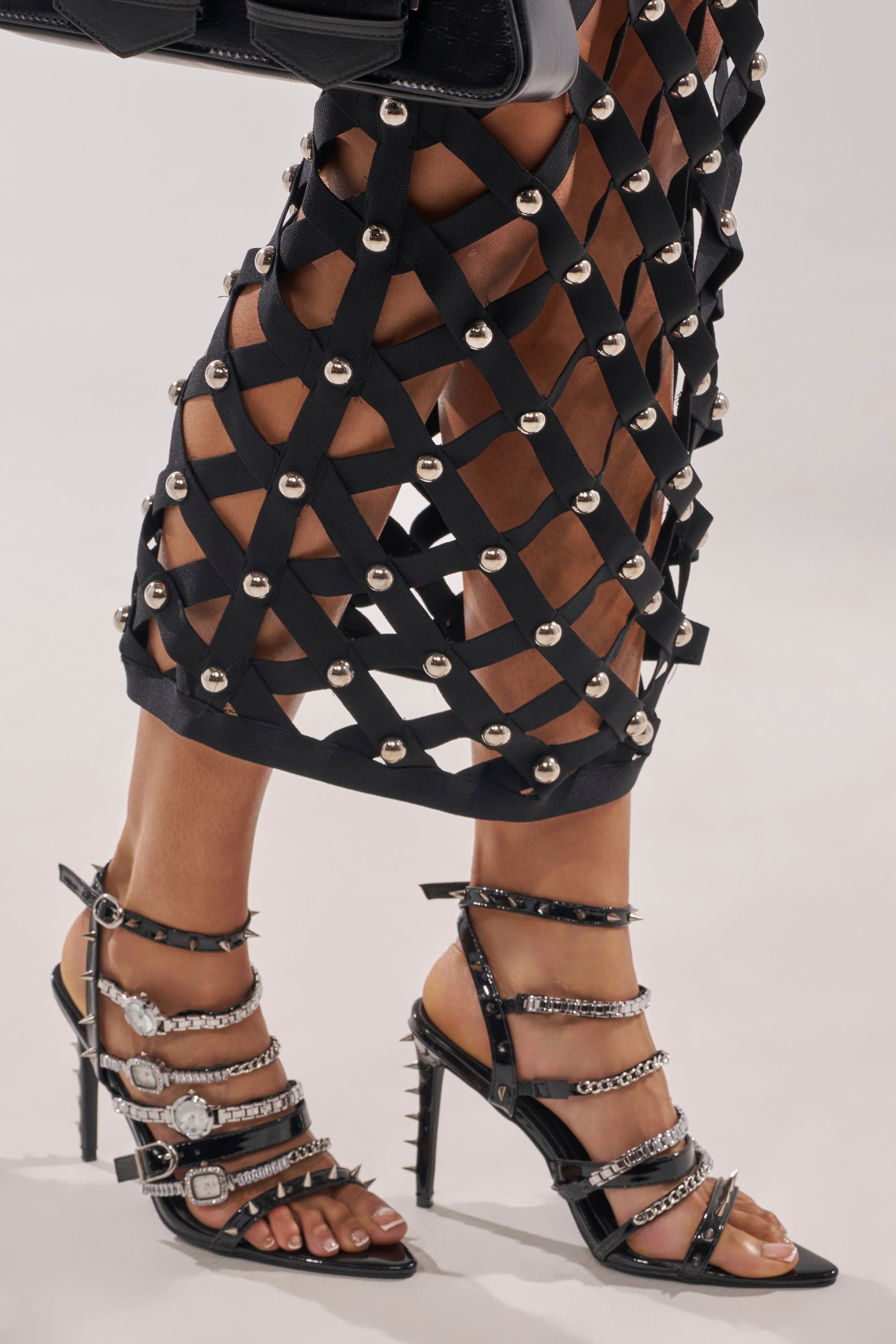 Wearing the AKIRA GOLD SYDONIC BLACK WATCH STILETTO SANDAL with silver accents and a black lattice skirt adorned with large circular studs, the person stands against a plain, light-colored background. 3