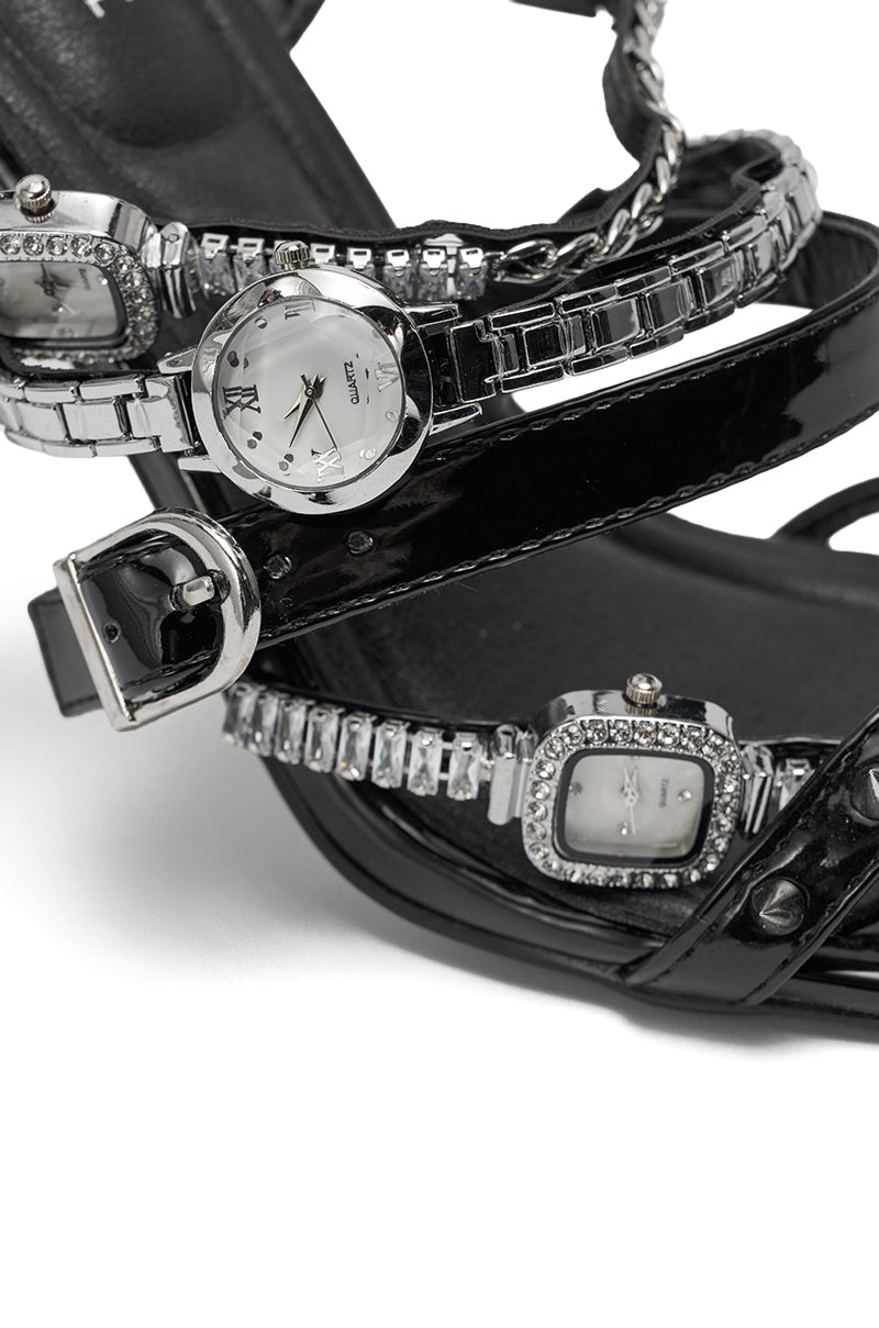 A close-up of the AKIRA GOLD SYDONIC BLACK WATCH STILETO SANDAL, featuring black shiny straps adorned with jeweled details and three visible watch faces, all set on a striking stiletto heel. 8