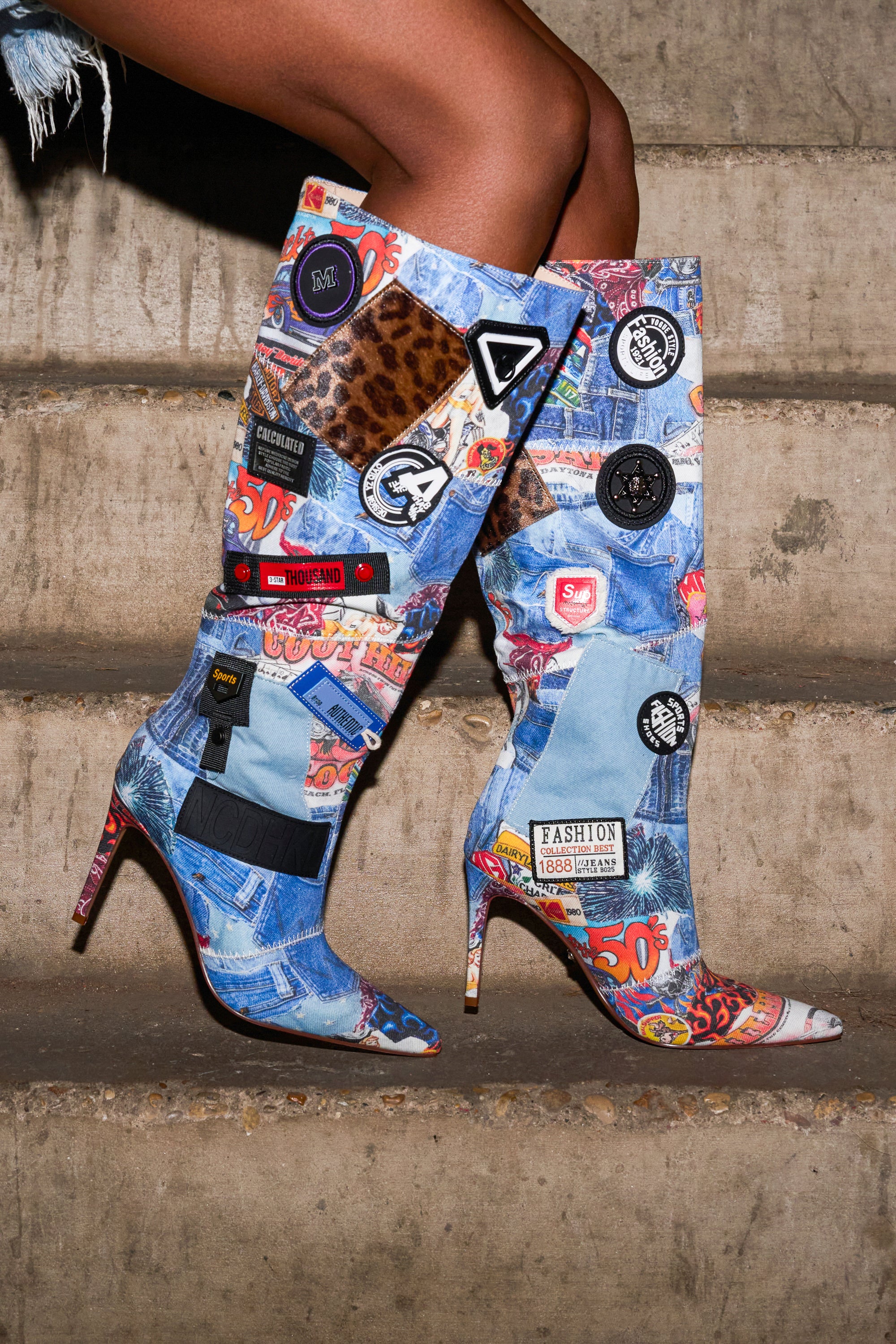 A person stands on concrete steps, wearing the AZALEA WANG SYLRILA MULTI ABSTRACT BOOT. Only their lower legs and these knee-high boots, featuring colorful denim patches, animal print, and badges in an abstract design, are visible.