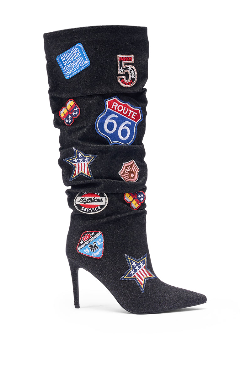 The AZALEA WANG SYLRILAAA DENIM MULTI BOOT is a black slouchy stiletto heel boot adorned with colorful embroidered patches like Route 66, American flags, and road signs, shown against a white background.