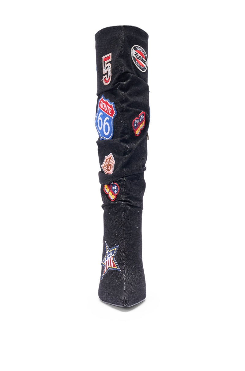 The AZALEA WANG SYLRILAAA DENIM MULTI BOOT features a tall black design with colorful embroidered patches like a Route 66 sign, star, and badges. Photographed from the front against a white background, these boots stand out.